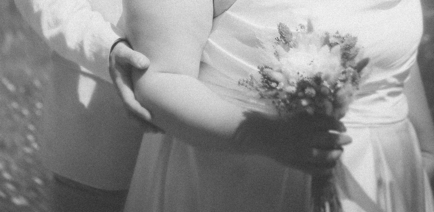 Black and white close-up of a bride holding a small floral bouquet with groom beside her.