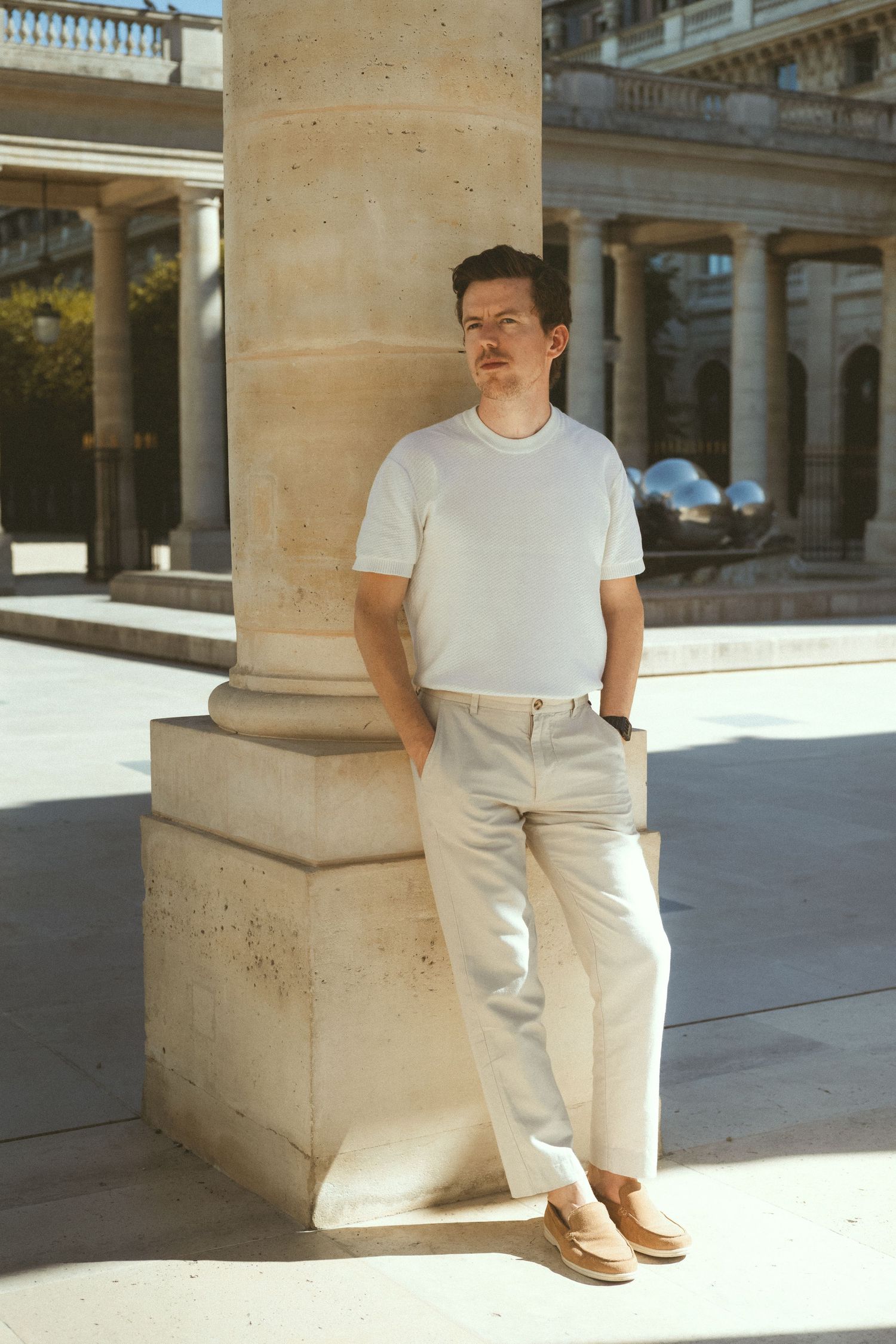 Man in white t-shirt and cream trousers leaning against a stone column in a sunlit classical courtyard.