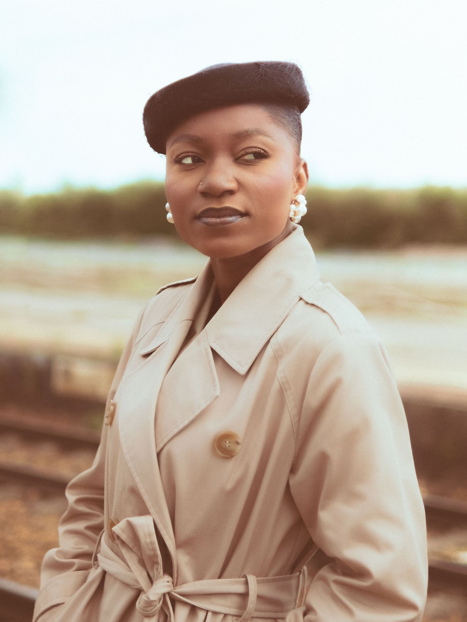 Woman in beige trench coat and black beret poses outdoors near railway tracks with blurred green background.