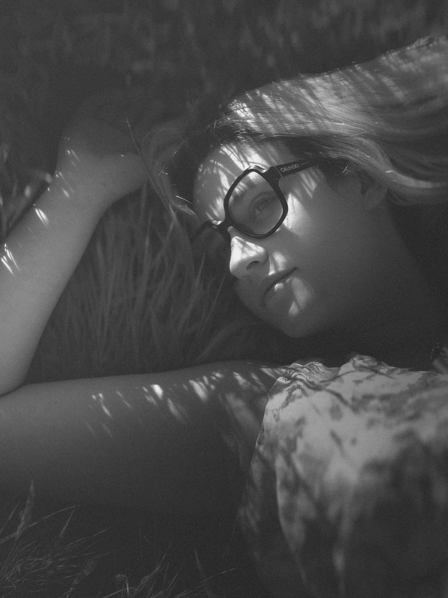 Black and white portrait of a young woman with glasses lying in grass, with dappled sunlight on her face.