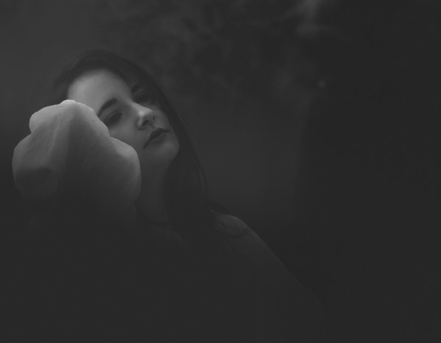 Black and white portrait of a young woman with closed eyes, resting her face against her hand in a moody, dark setting.