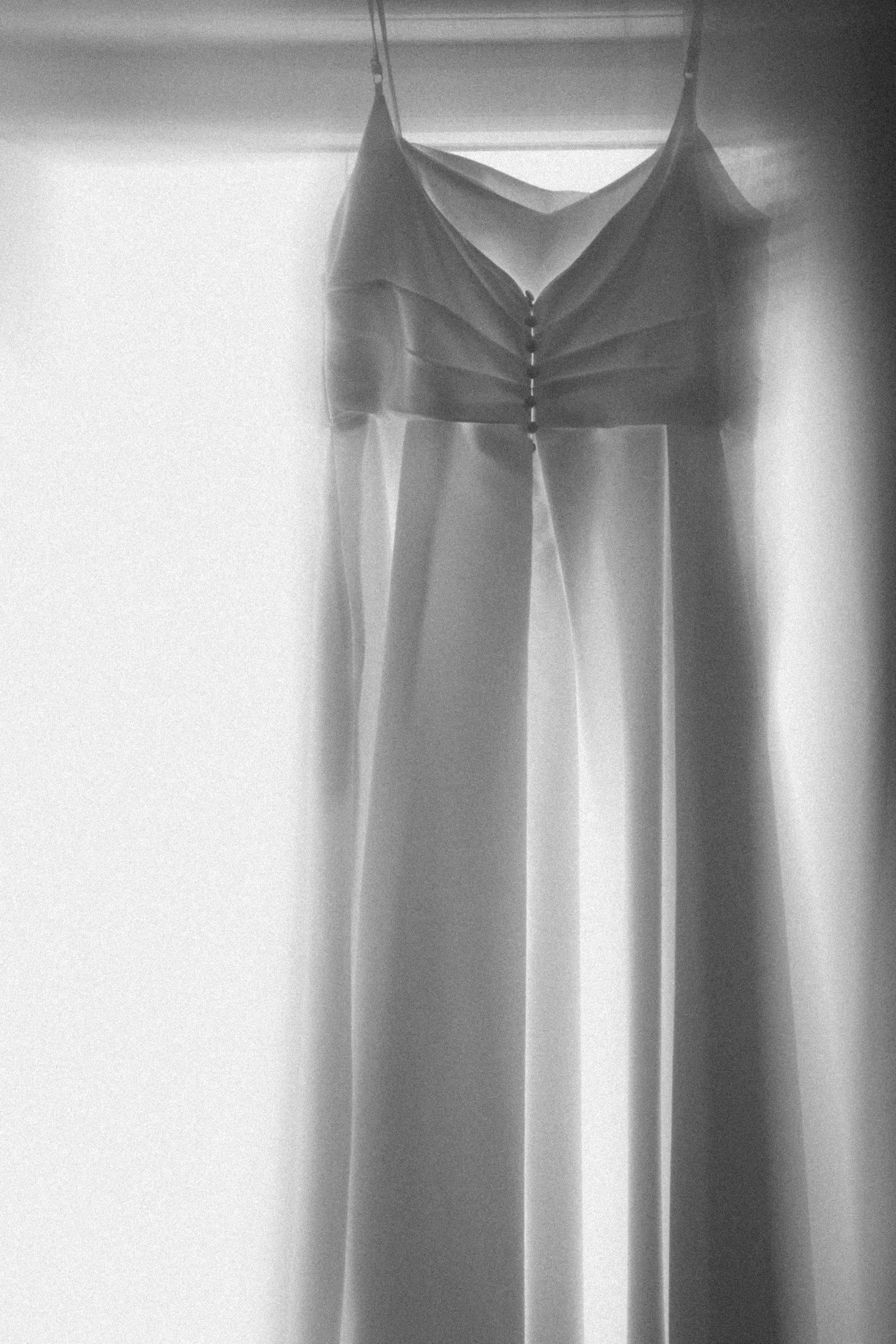 Photo élégante en noir et blanc d'une robe de mariée suspendue près d'une fenêtre ensoleillée.