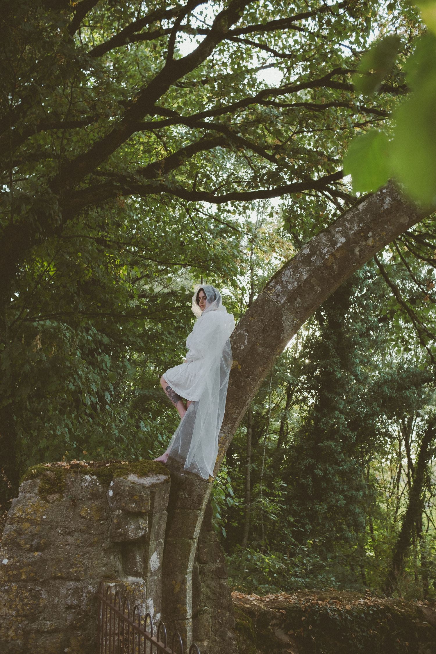Bride in white gown and veil stands atop ancient stone arch surrounded by lush green forest trees.