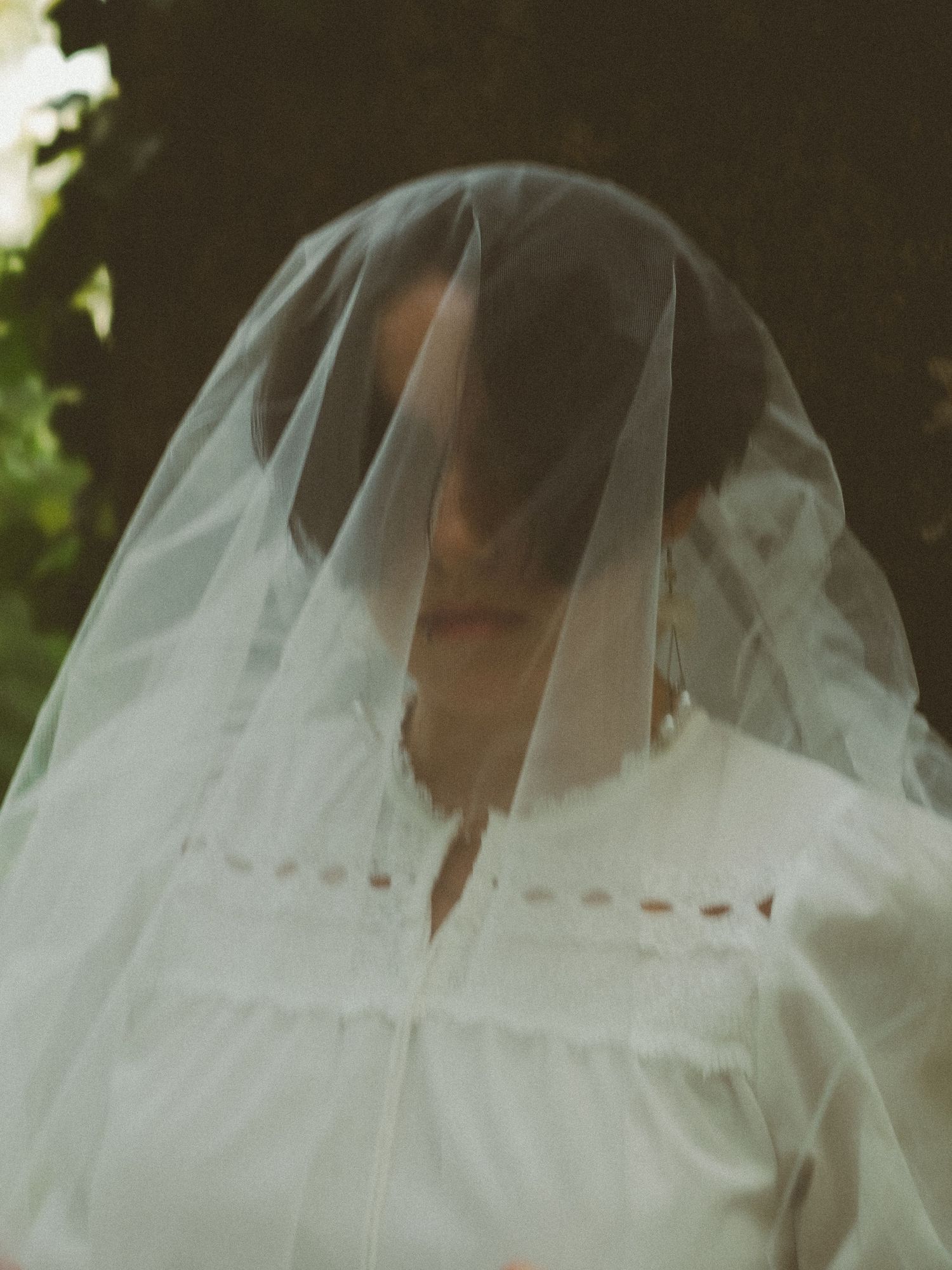 A bride wearing a white dress and sheer veil draped over her head, standing outdoors near green trees.