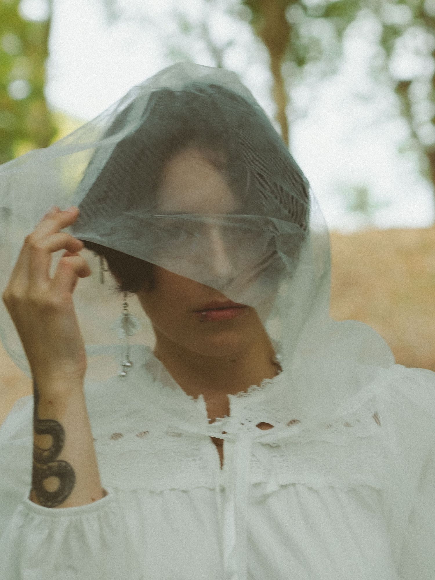 Woman in white eyelet dress holds sheer fabric over face revealing tattoo on wrist.