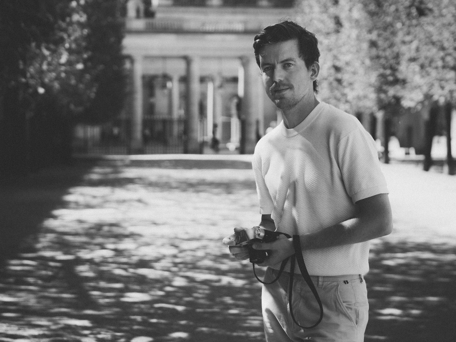Black and white photo of a young man holding a camera outdoors in a sunlit park.