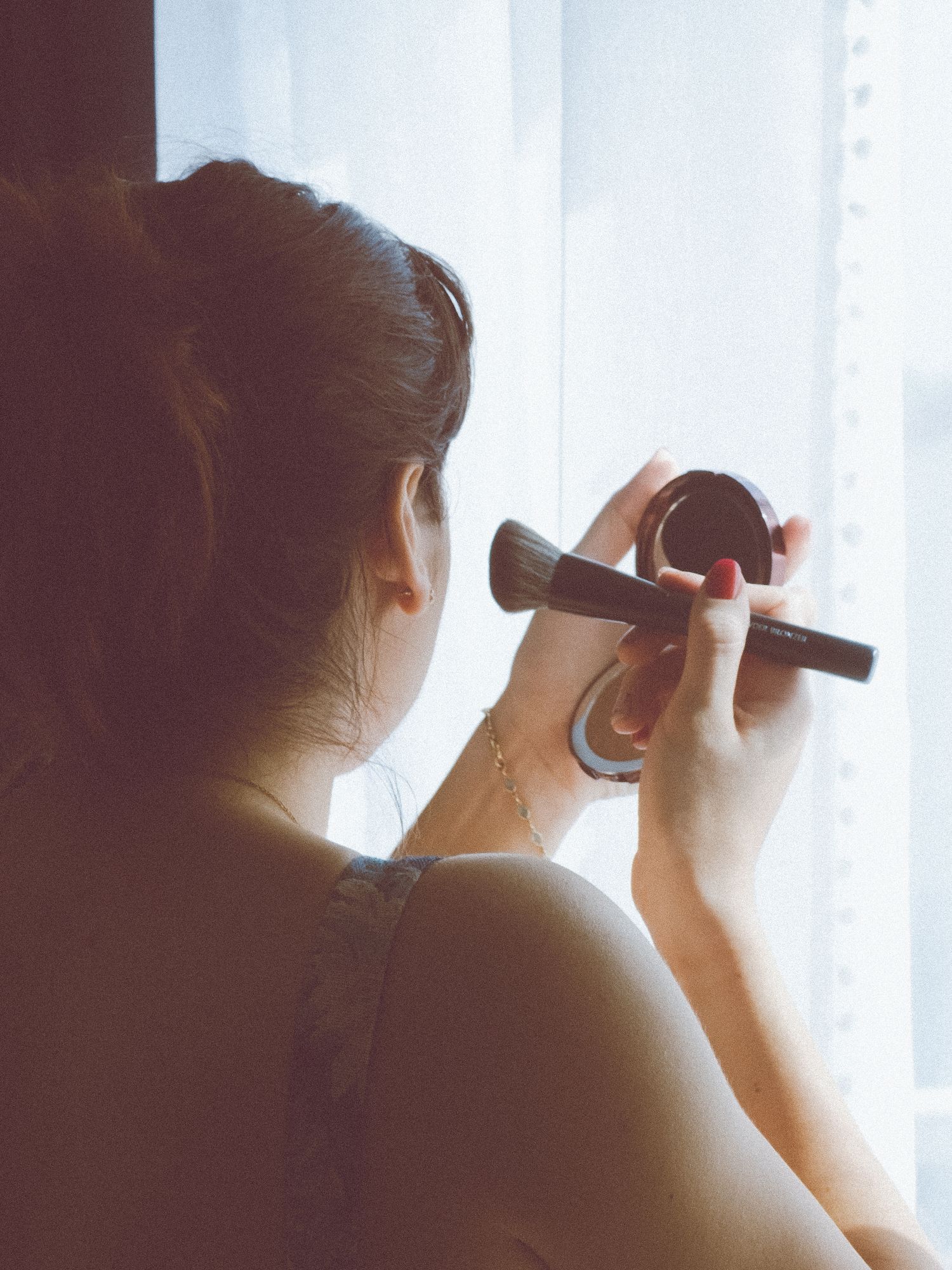 Femme se maquillant avec un pinceau et un miroir compact, assise près d'une fenêtre lumineuse.