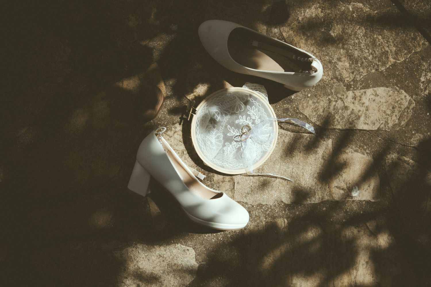 White bridal heels and a round lace clutch purse arranged on stone ground with dappled sunlight and shadows.
