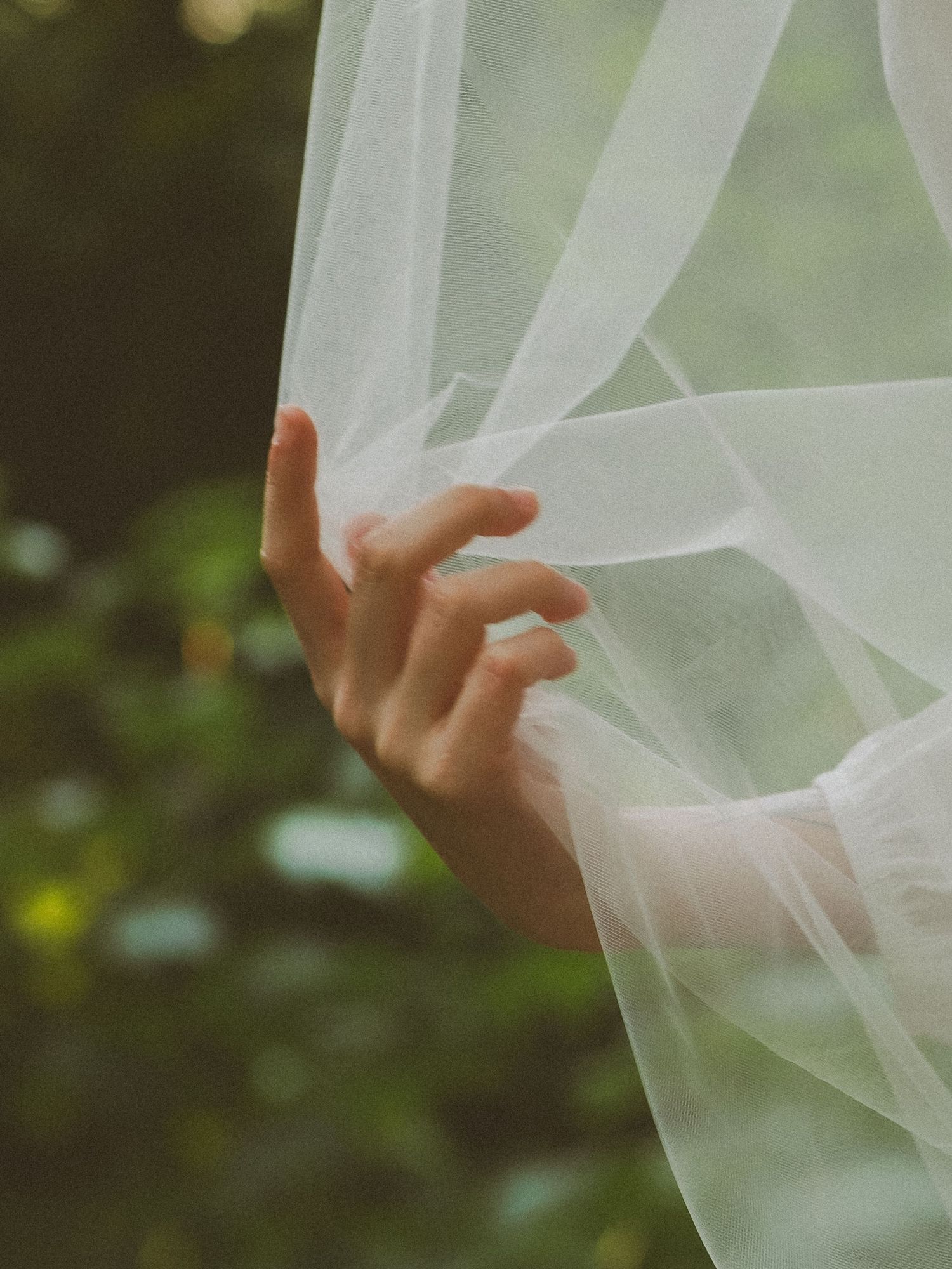 A delicate hand gently holds sheer white tulle fabric outdoors with lush green foliage in the background.