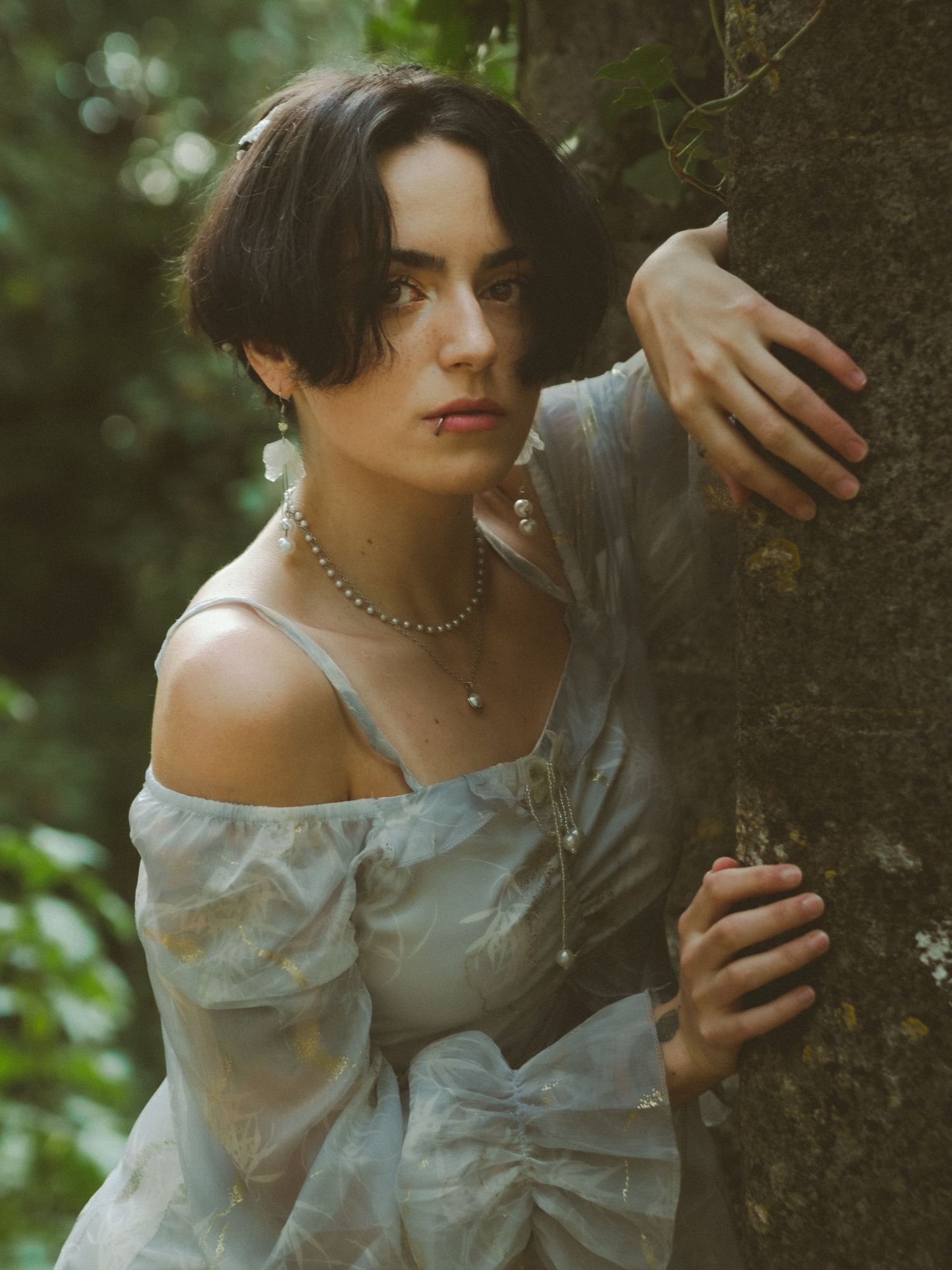 Dark-haired woman in blue off-shoulder dress with pearl necklace leans against tree in forest.
