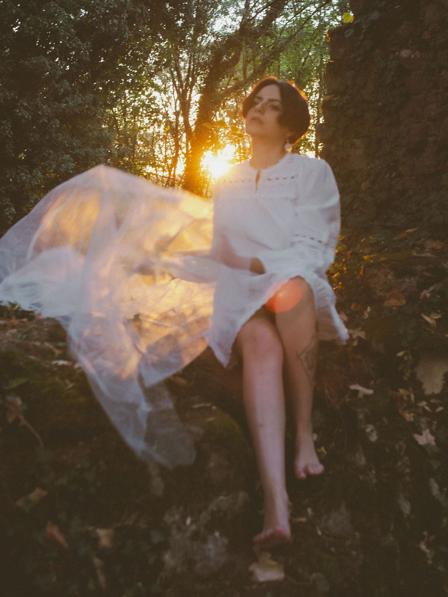 Barefoot woman in white dress sits on rocks at golden sunset in forest.