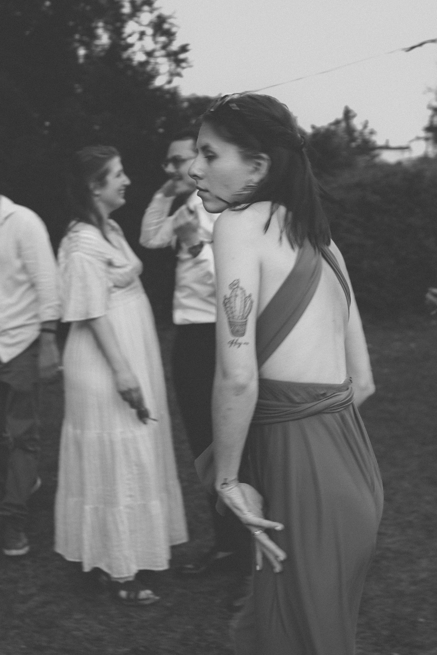 Black and white photo of a woman with a tattoo in a backless dress at an outdoor gathering.