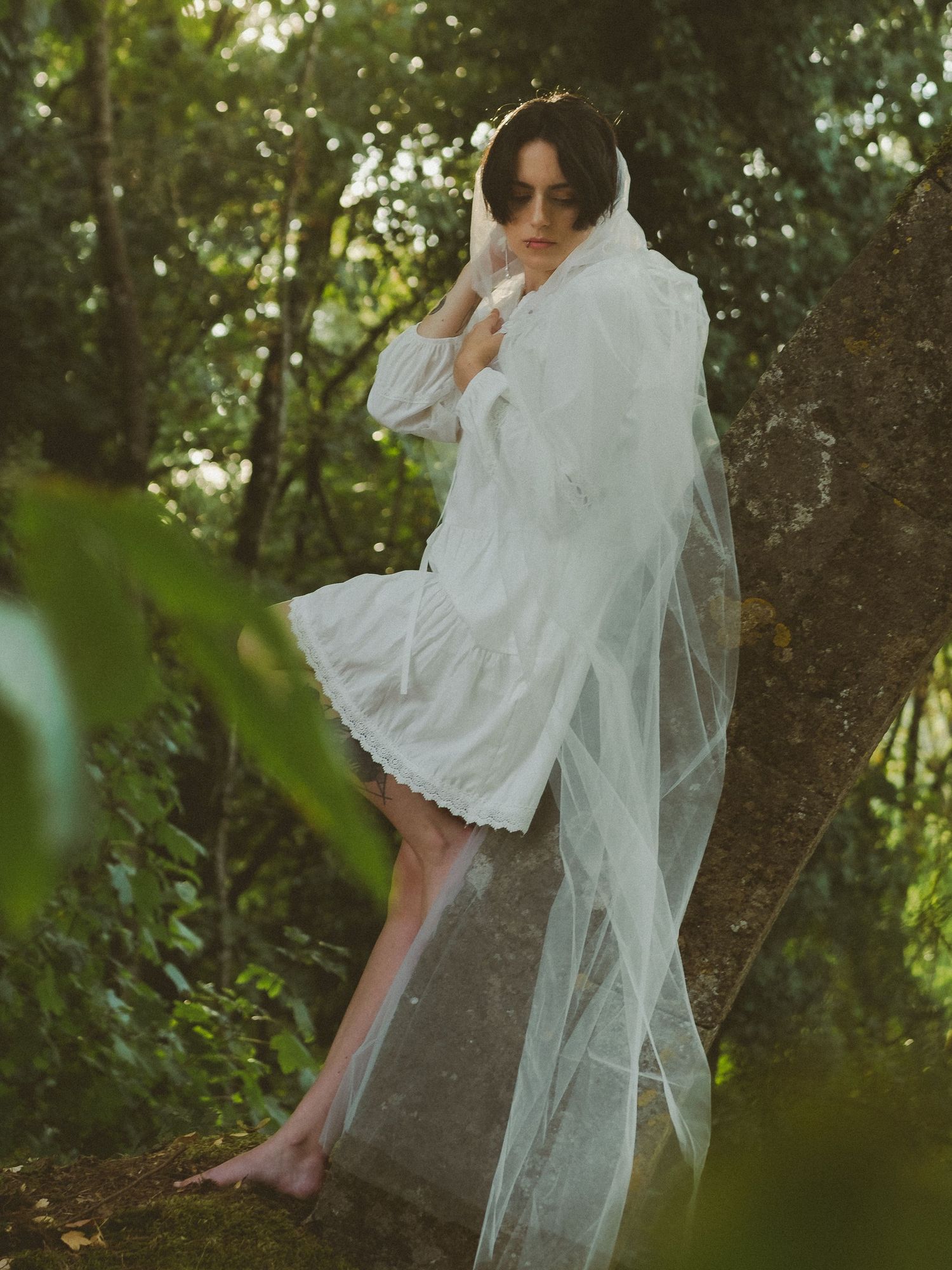 Barefoot woman in white lace dress with tulle veil sits on stone in forest.