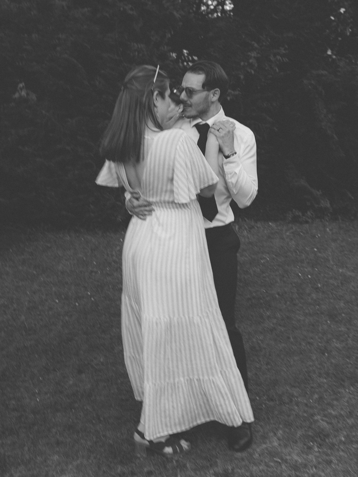 Photo noir et blanc d'un couple partageant un baiser romantique tout en dansant sur une pelouse en extérieur.