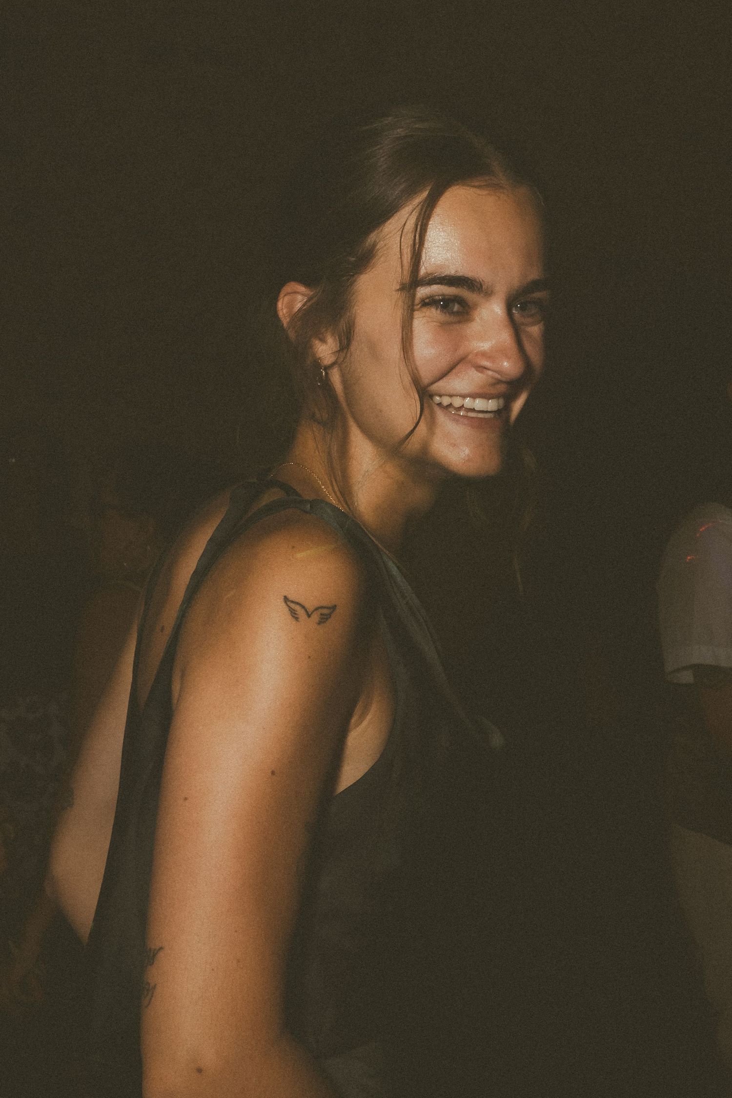 Smiling young woman with a small butterfly tattoo on her shoulder wearing a black tank top in dim lighting.