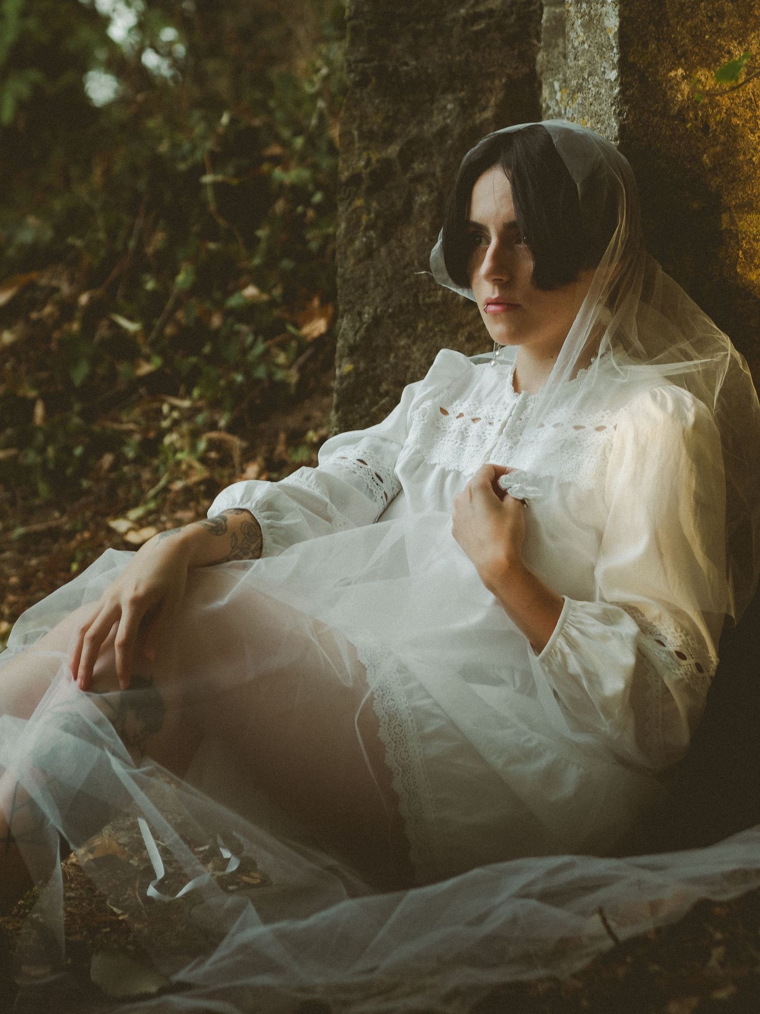 A woman in a white dress and veil sits pensively against a mossy tree in a dreamy outdoor setting.