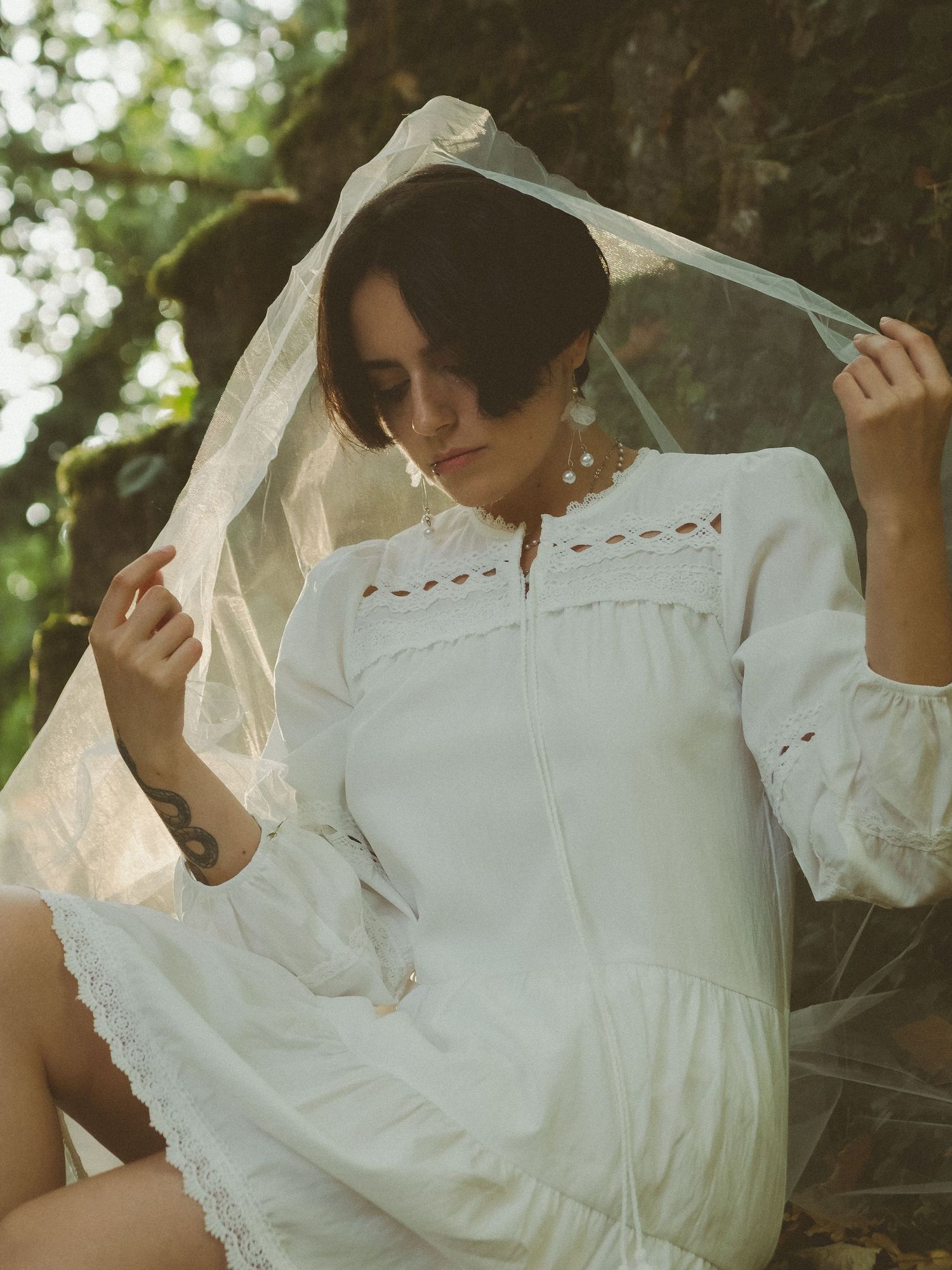 Woman in white embroidered dress holding sheer veil over head in forest setting.