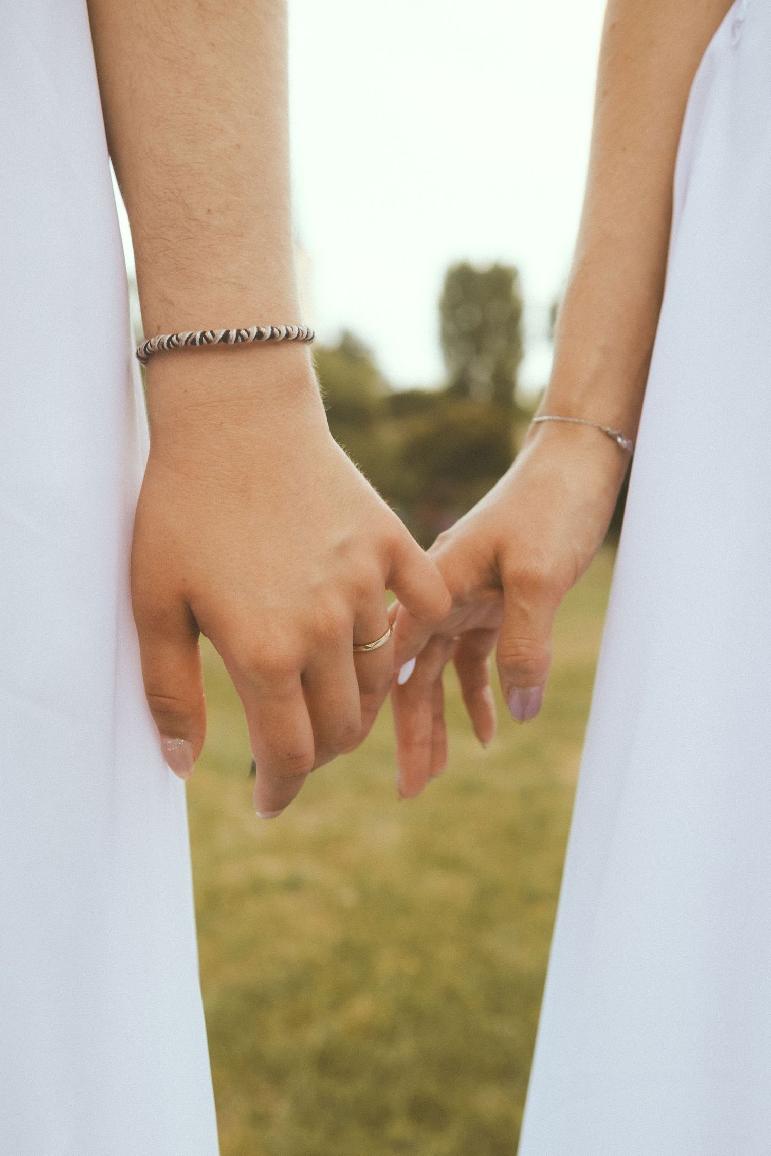 Two people holding hands with bracelets and a ring, dressed in white, standing in a green field.