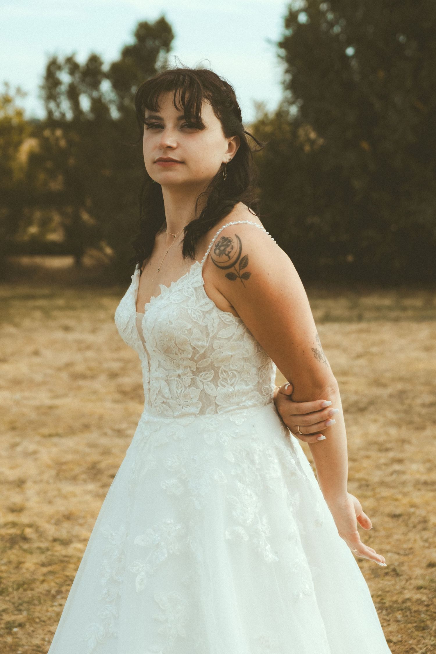 Bride in white lace A-line gown with shoulder tattoo posing outdoors in golden light.