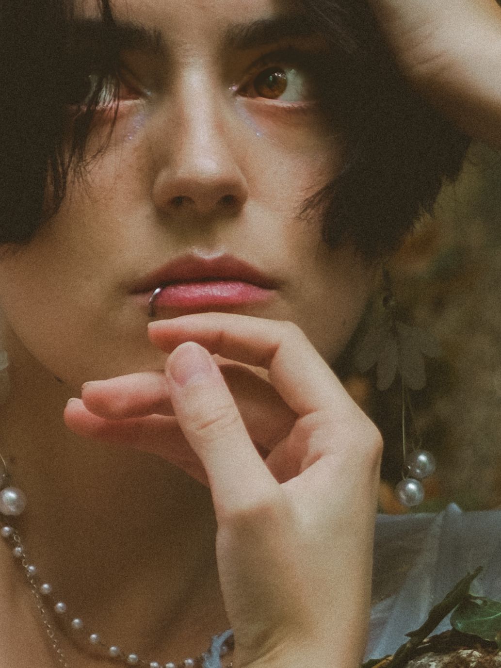Close-up portrait of a young person with dark hair, pearl necklace, and earrings, resting hand on chin.