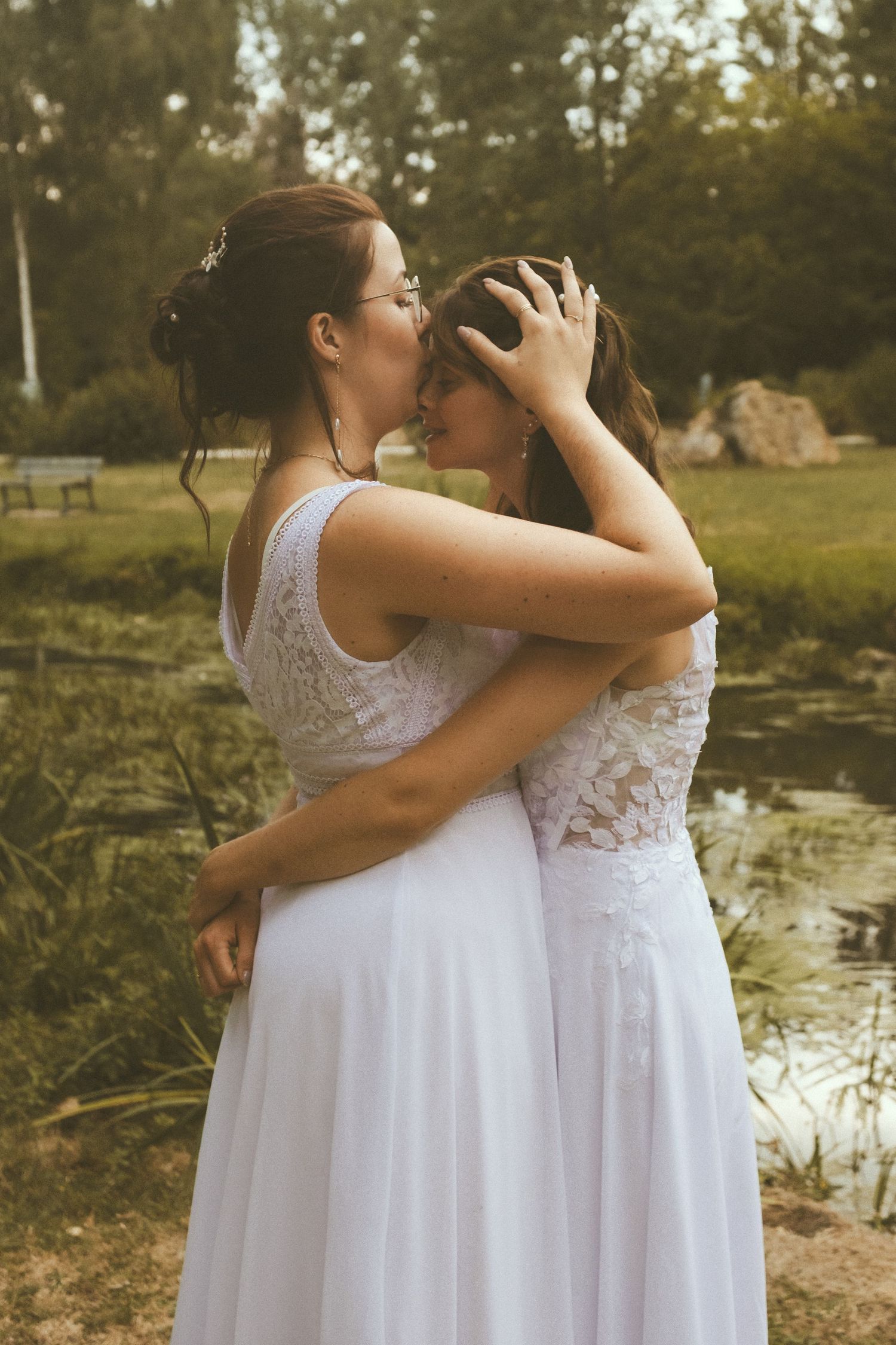 Two brides in white lace gowns share a tender kiss outdoors near a pond surrounded by lush greenery.