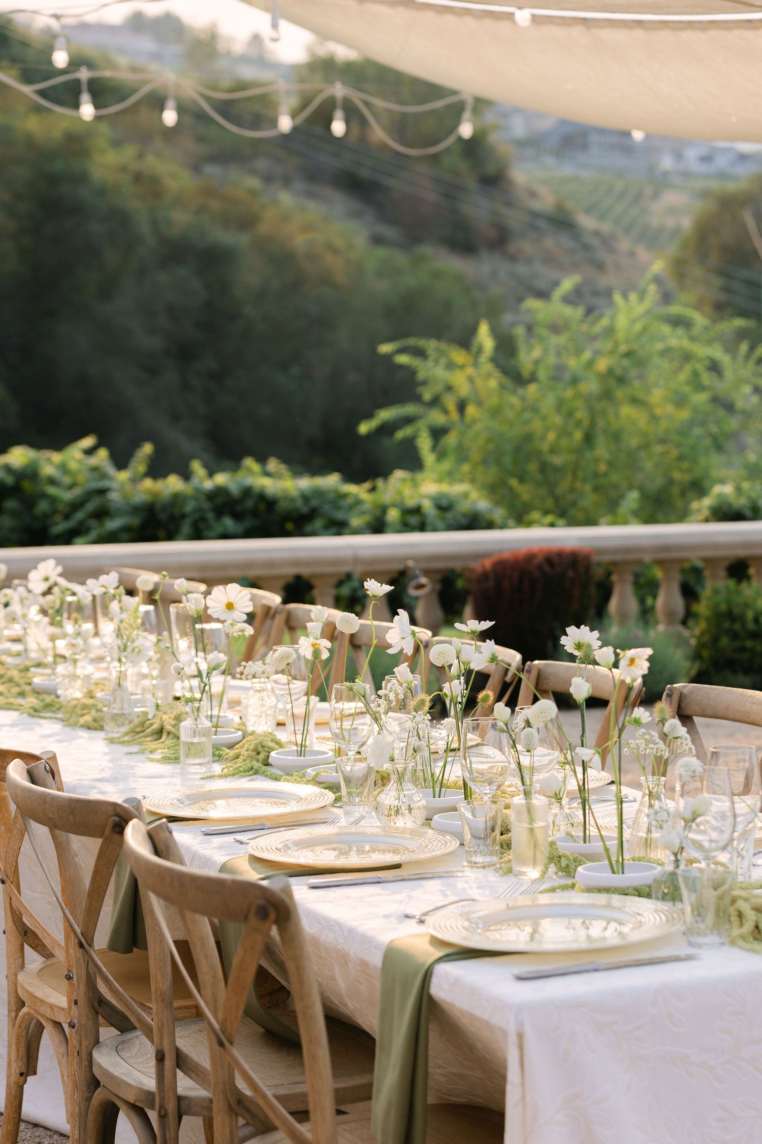 Elegant outdoor wedding reception table setting with delicate floral arrangements and rustic wooden chairs.