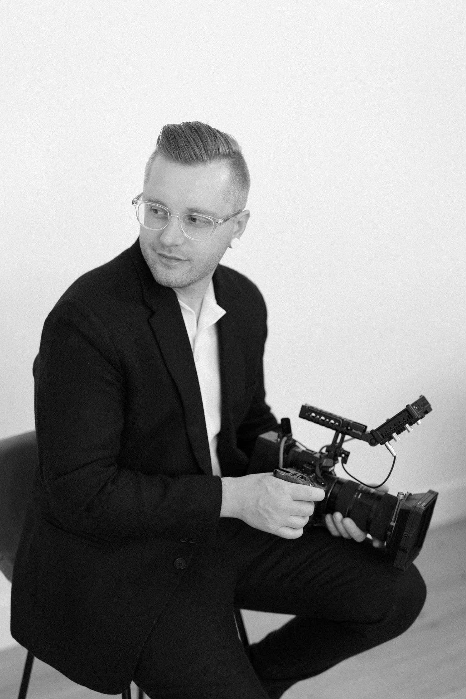 Black and white photo of a man in a suit holding a professional video camera, sitting and smiling at the camera.