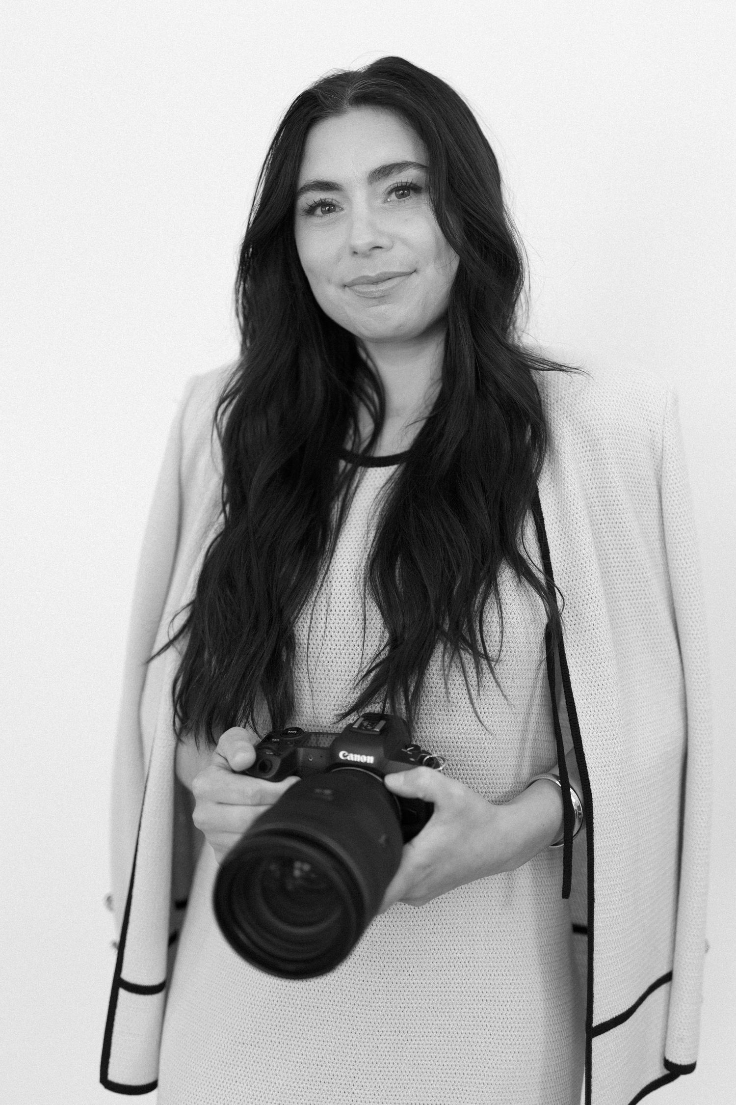 Black and white portrait of a woman with long dark hair holding a Canon DSLR camera, wearing a light blazer.