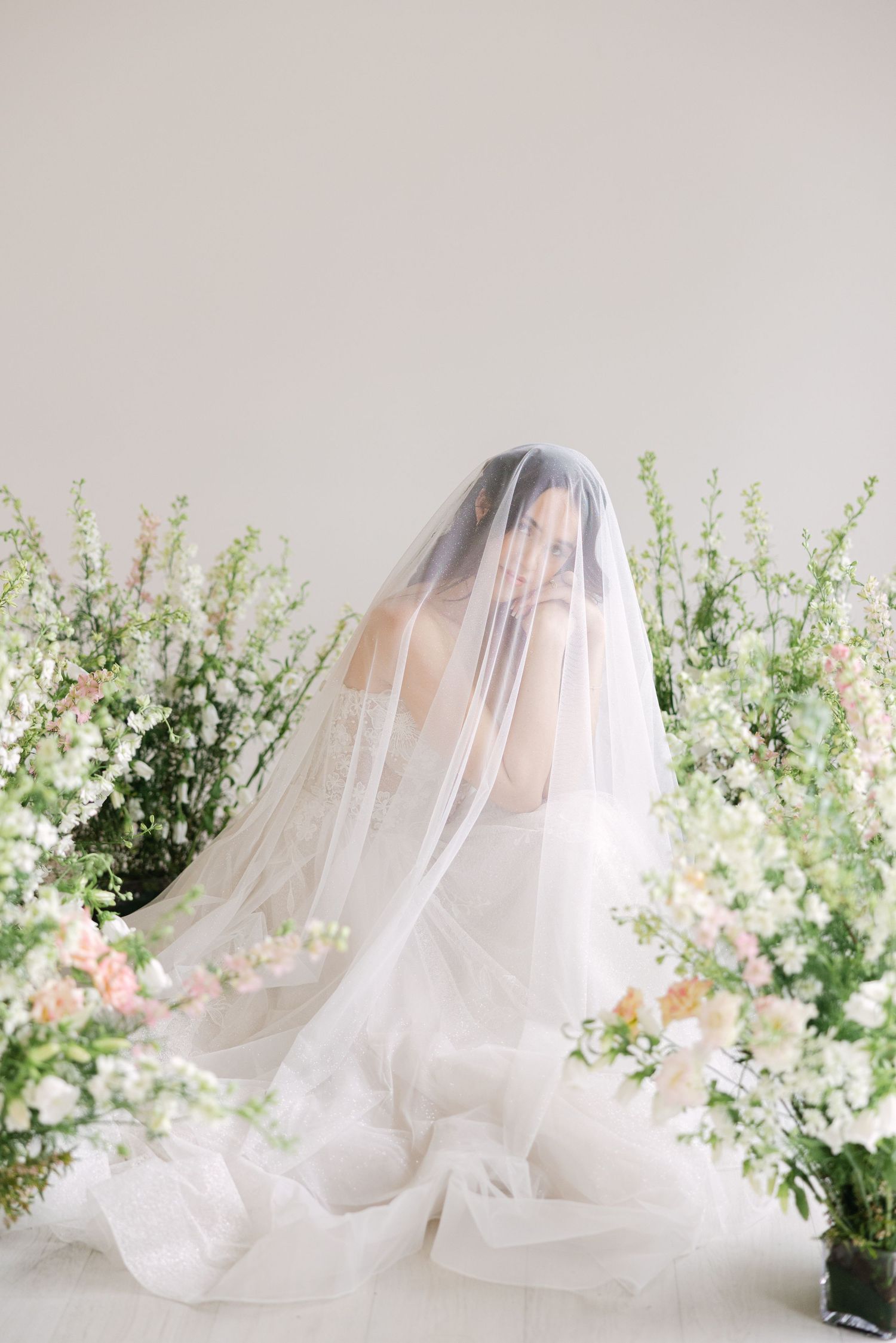 Ethereal bridal portrait with flowing veil surrounded by delicate white and pink floral arrangements.
