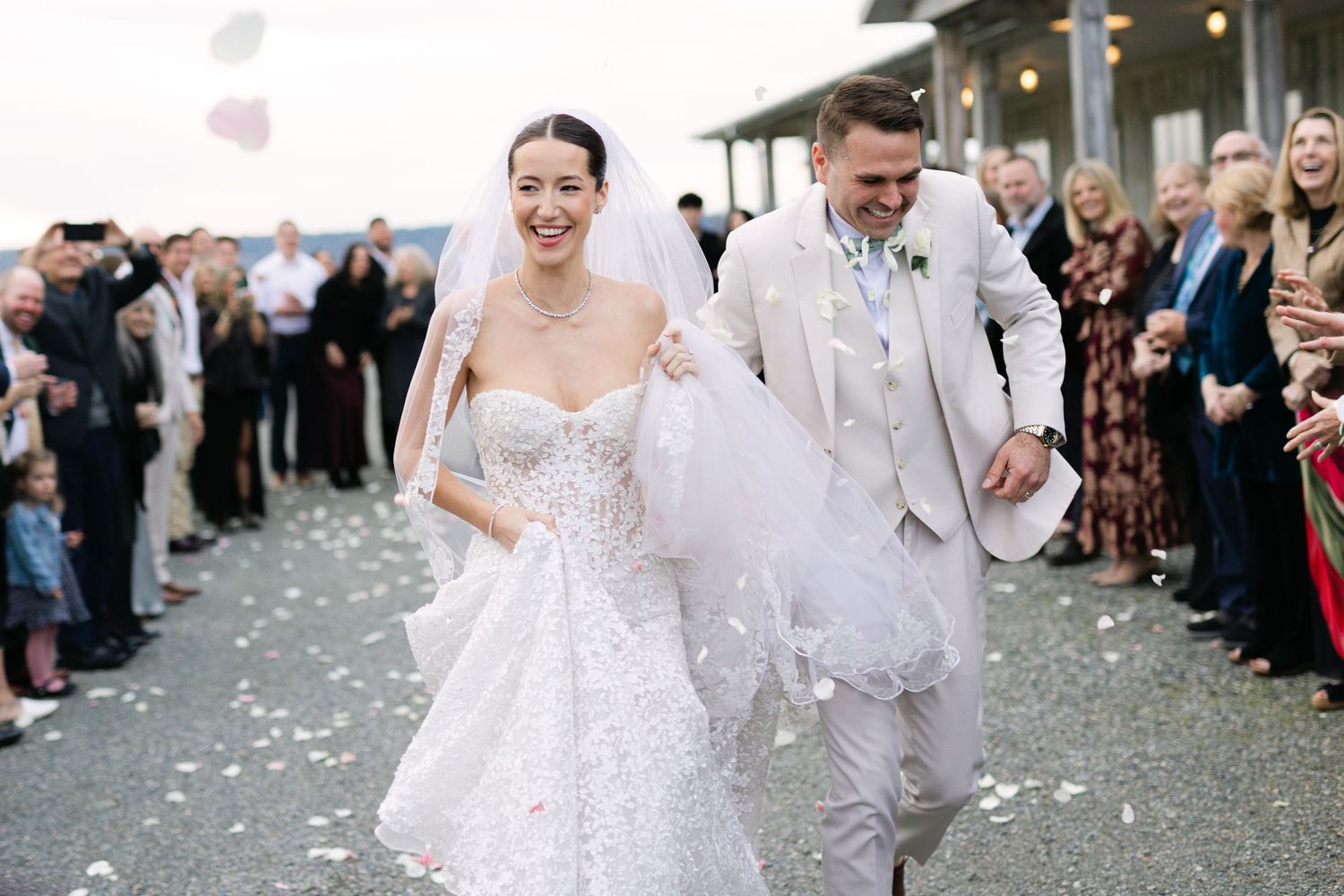 Newlyweds walk through shower of white confetti as wedding guests cheer along pathway.
