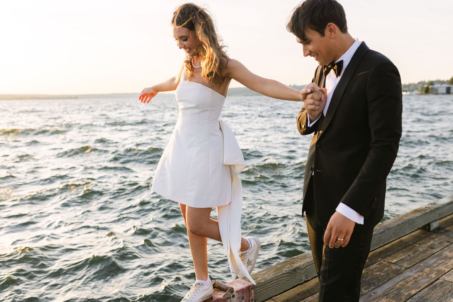 Couple in wedding attire walking along oceanfront boardwalk at sunset.