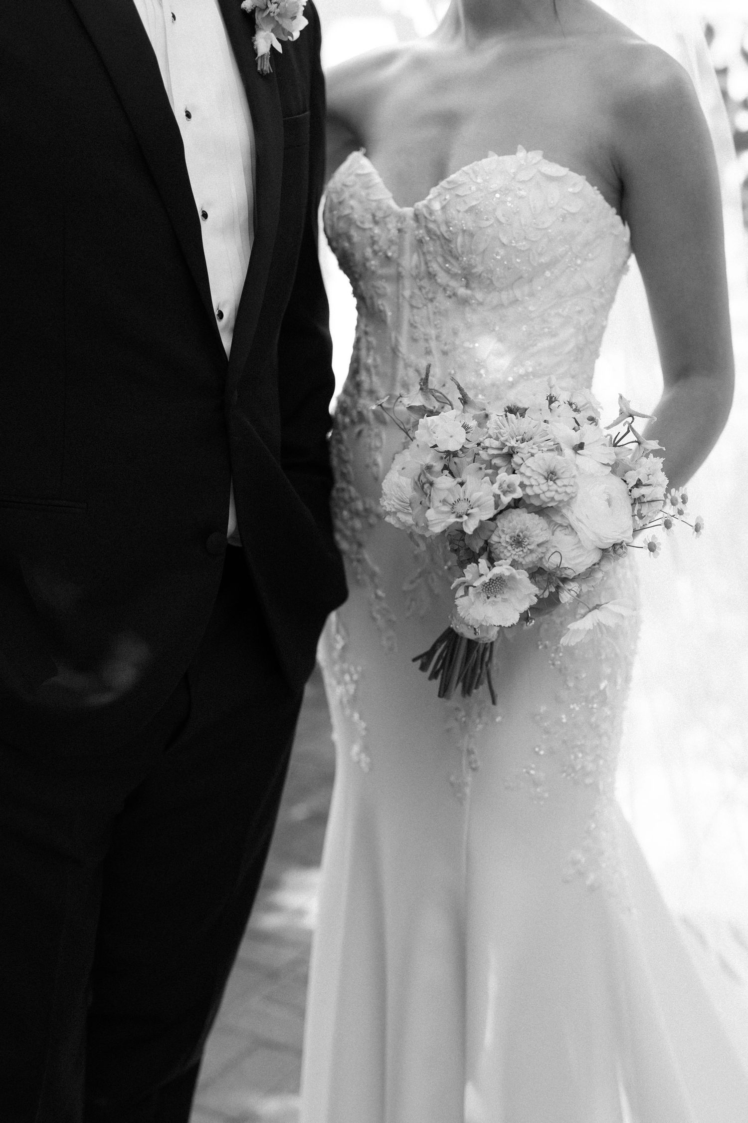 Black and white close-up of bride in lace dress with bouquet standing next to groom in tuxedo.
