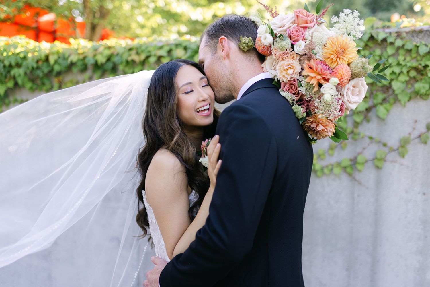 A couple in wedding attire share a romantic moment outdoors with a flowing veil and colorful floral bouquet.