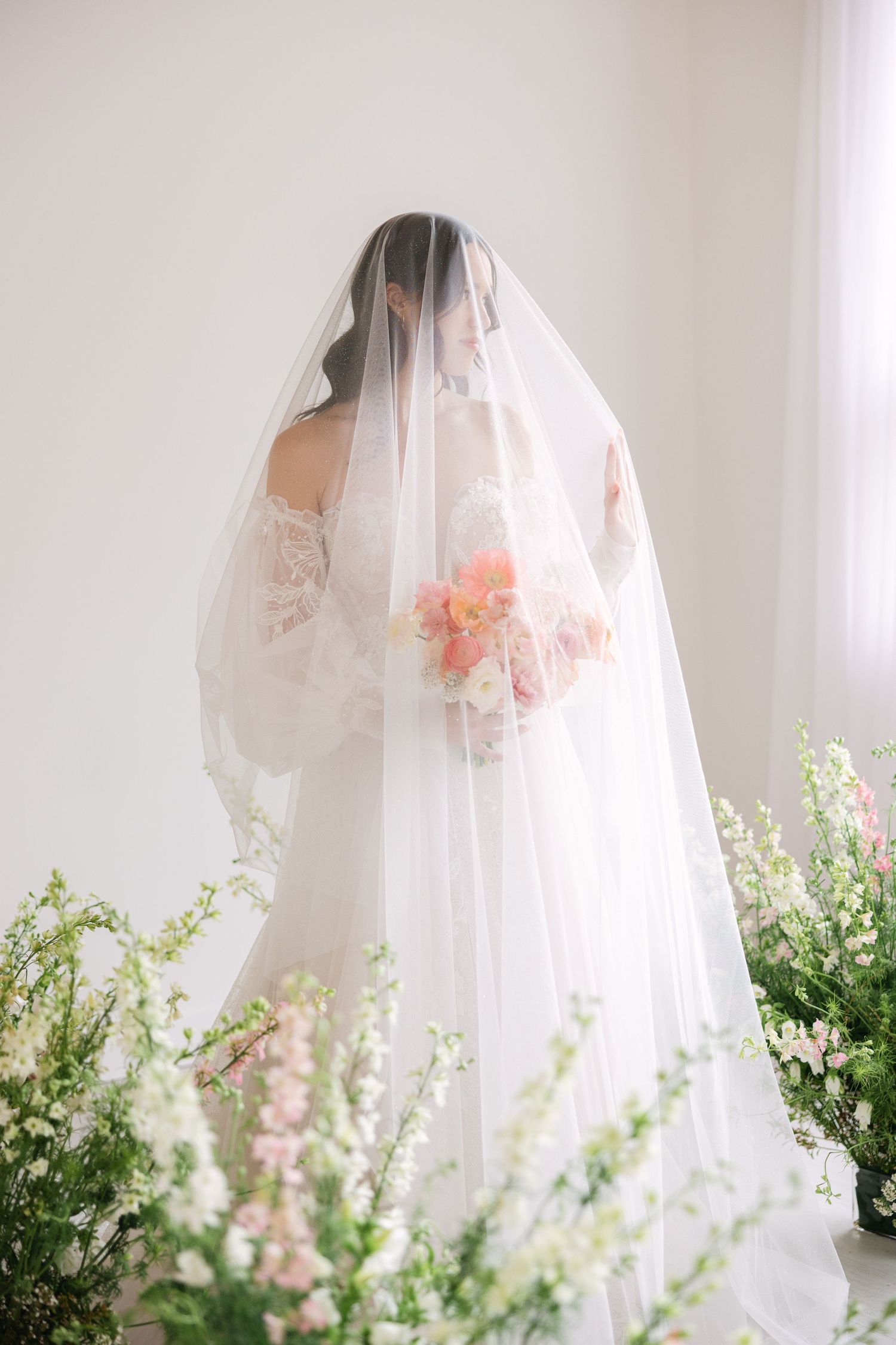Delicate bridal veil detail shot with soft pink flowers and greenery in the background.