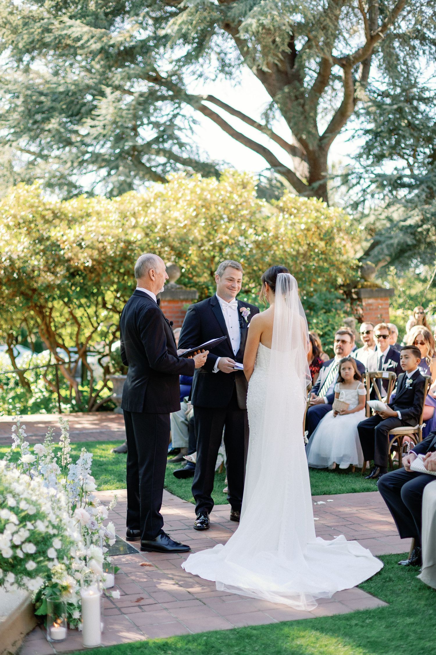 Outdoor garden wedding ceremony with couple exchanging vows under large tree.