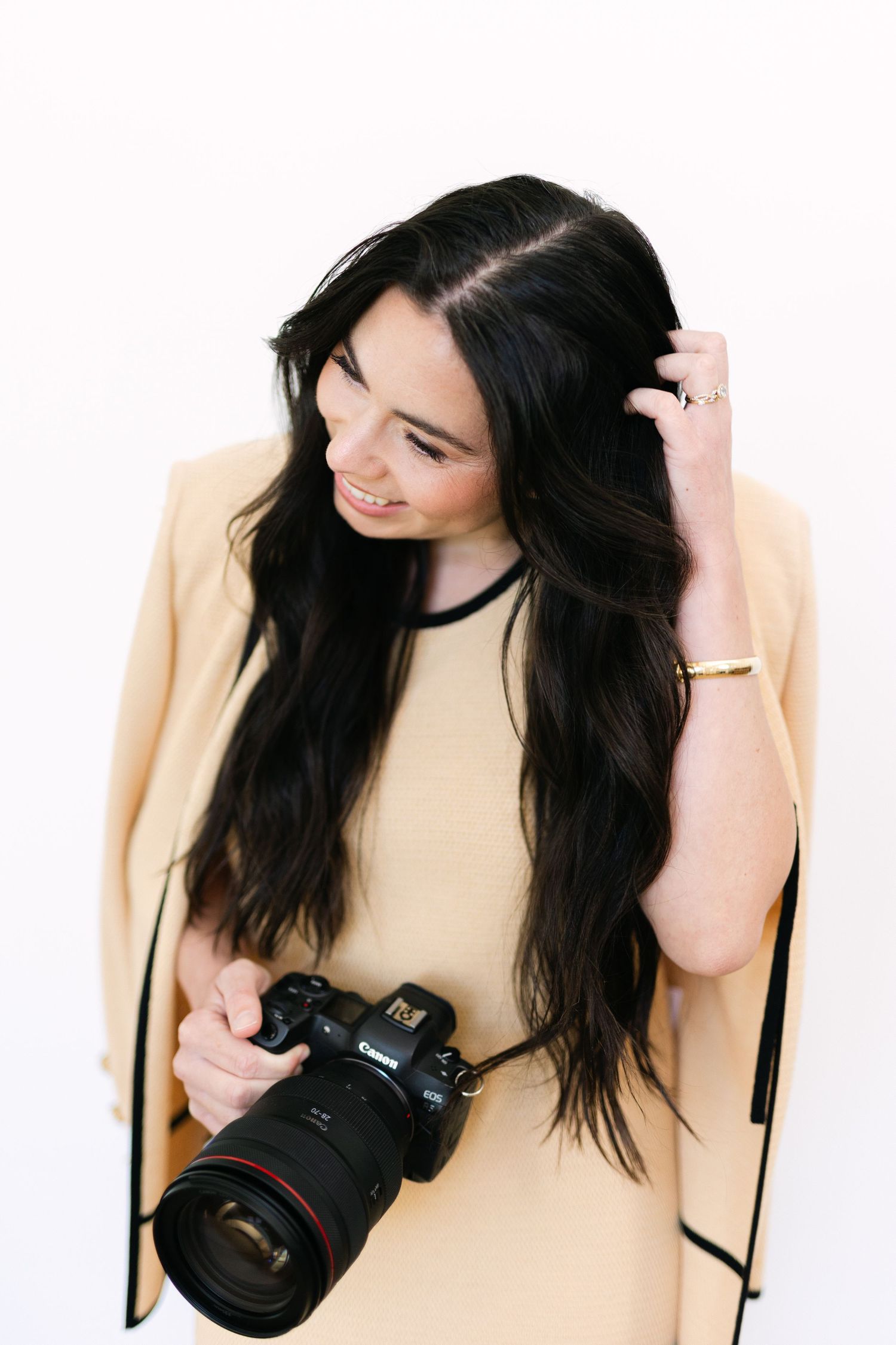 Smiling brunette woman holding a Canon DSLR camera, wearing a yellow outfit against a white background.
