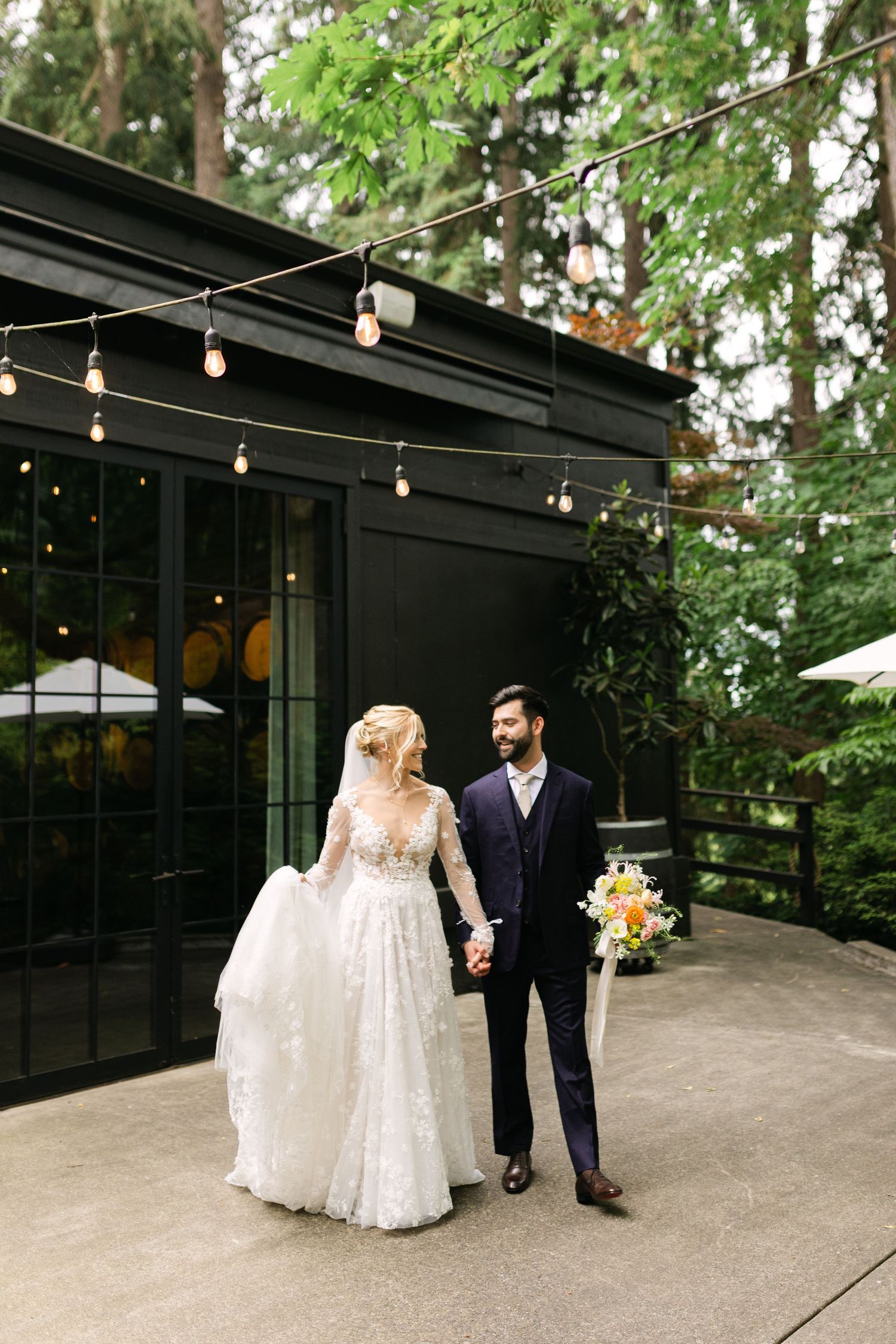 Couple walking outside modern venue with string lights and greenery overhead.