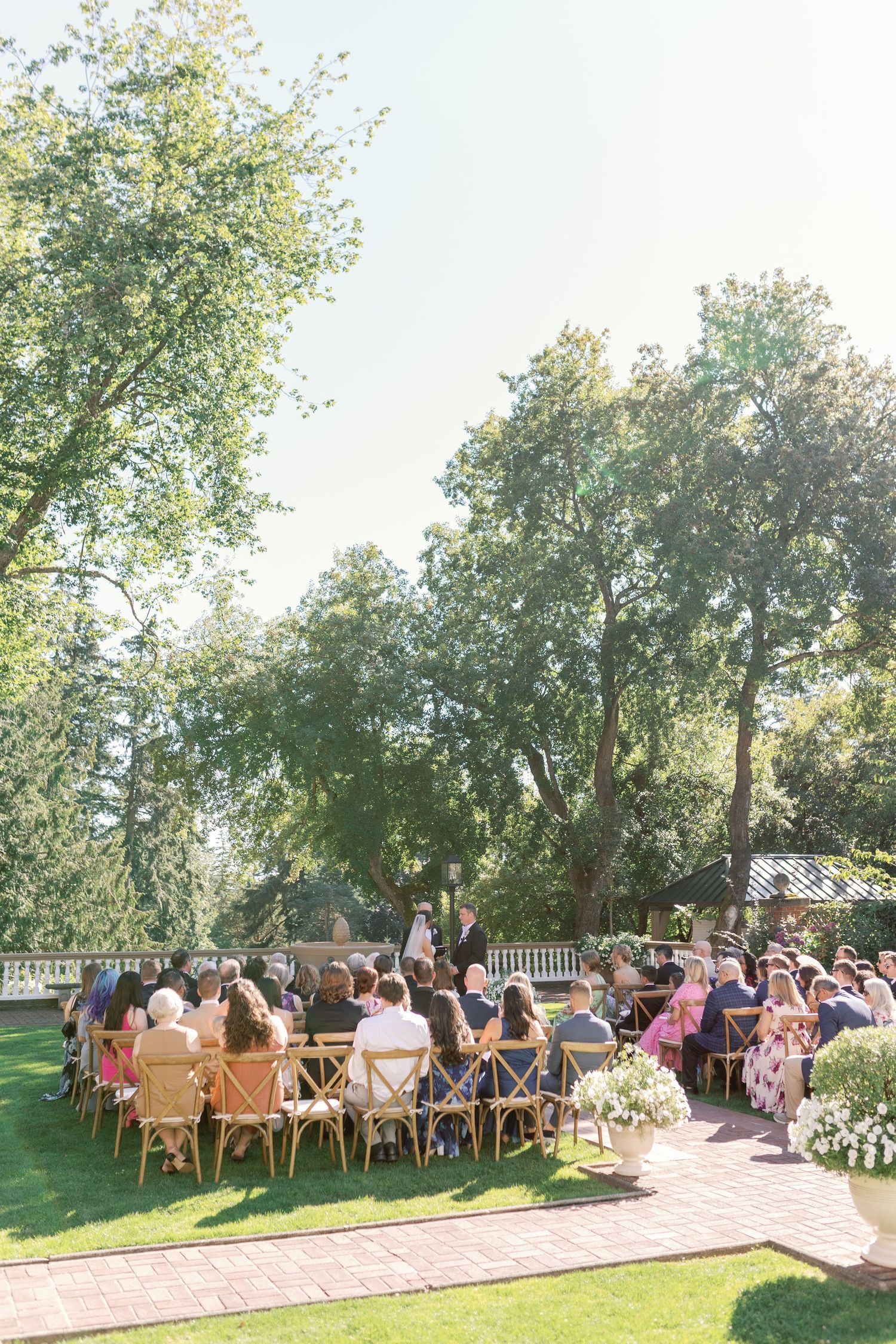 Wedding guests seated outdoors on wooden chairs beneath tall trees during an elegant garden ceremony.