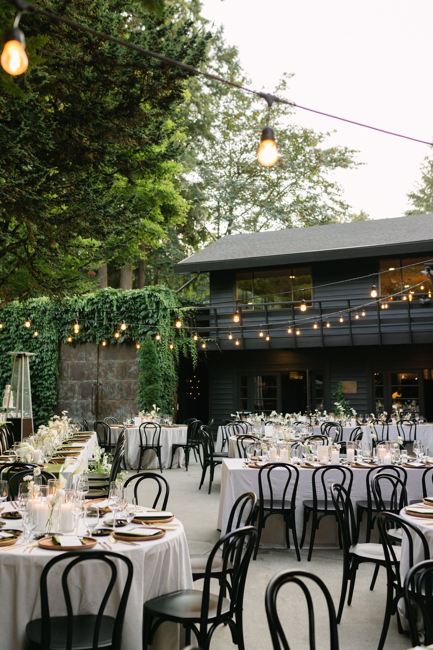 Outdoor wedding reception setup with string lights and black bentwood chairs under trees.
