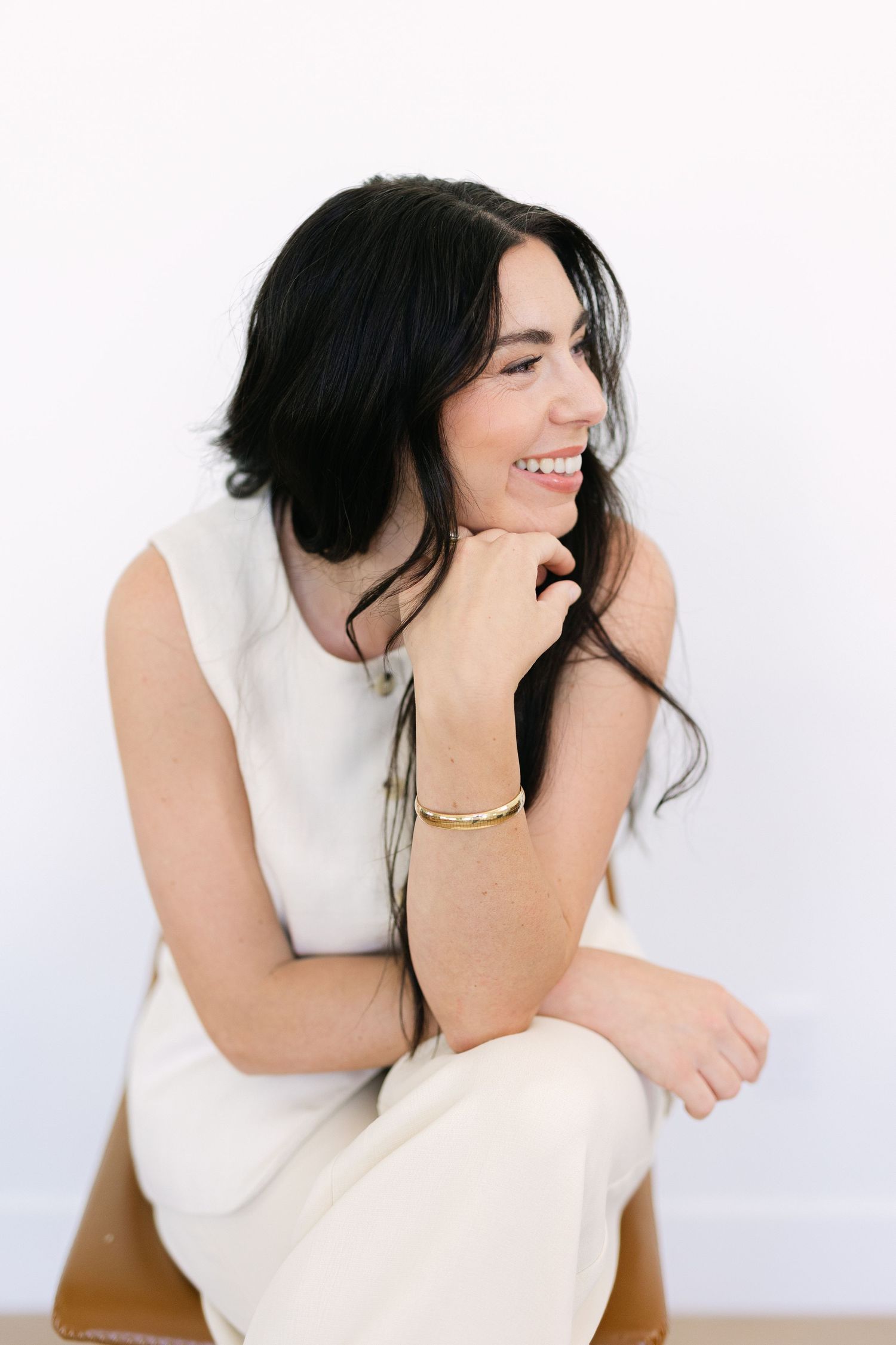 Smiling woman with dark hair wearing a white sleeveless dress and gold bracelet, sitting relaxed against a white background.