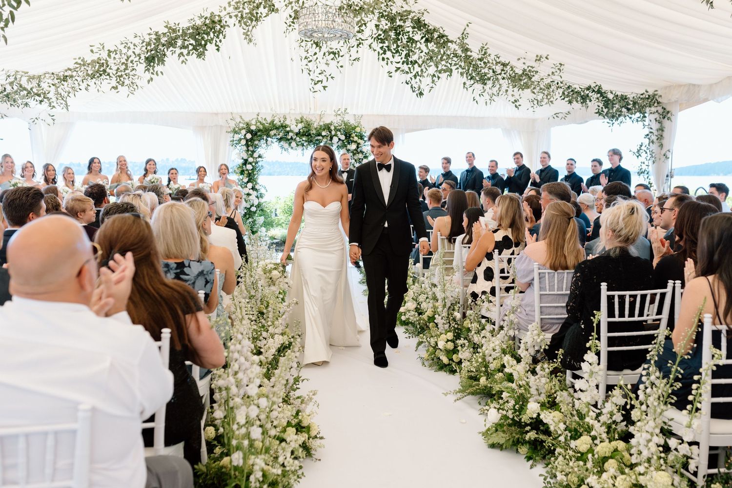 Bride and groom walk down floral-lined aisle in elegant white tent wedding ceremony with lush greenery arch.