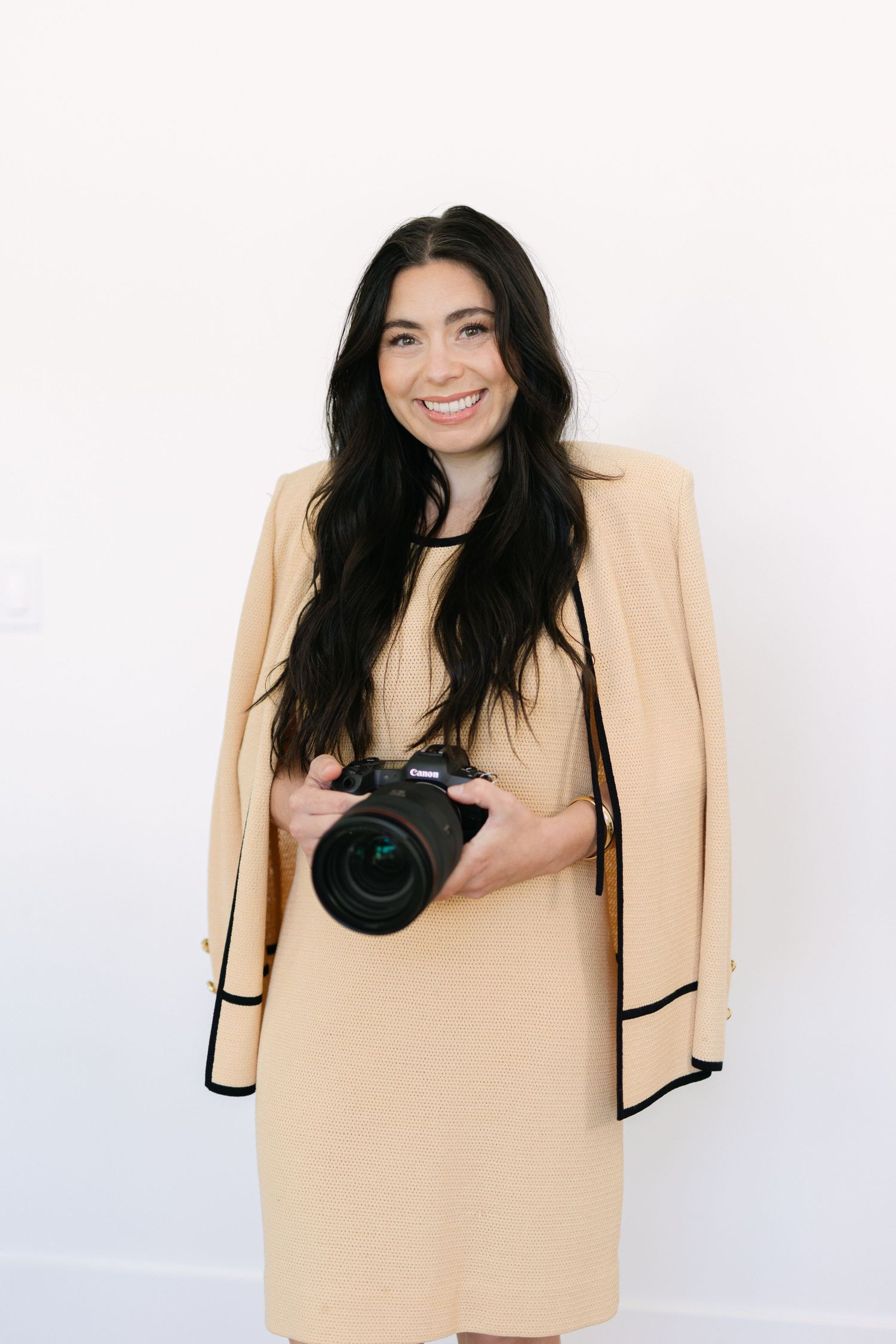 Smiling woman with long dark hair holding a DSLR camera, wearing a beige dress and blazer against a white background.