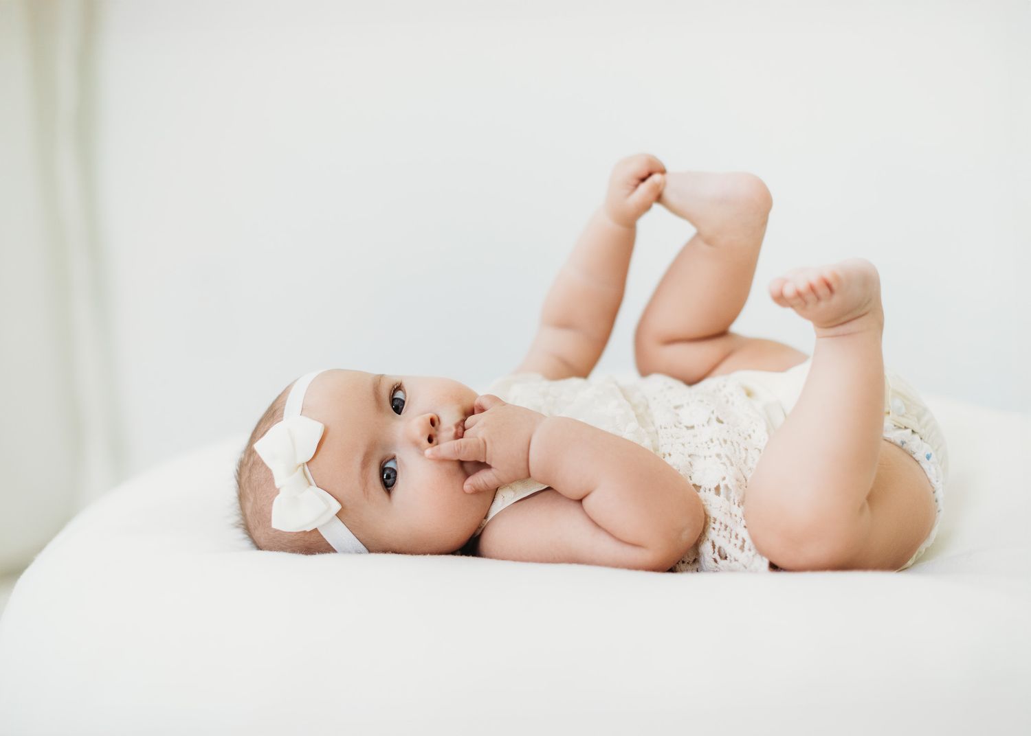 Baby in white onesie lying playfully on white bedding during photoshoot.