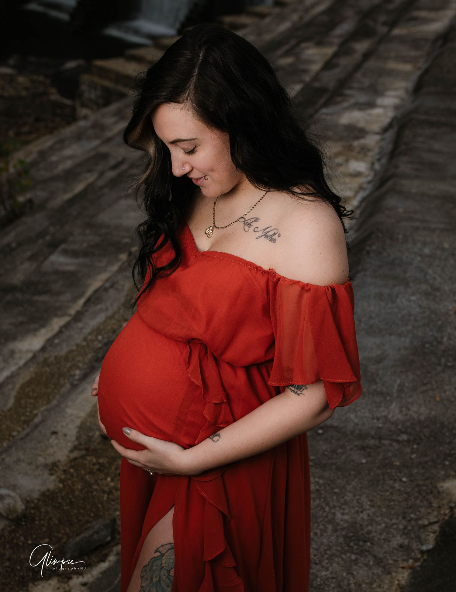 Pregnant woman in flowing red off-shoulder dress tenderly holding baby bump during outdoor maternity session.