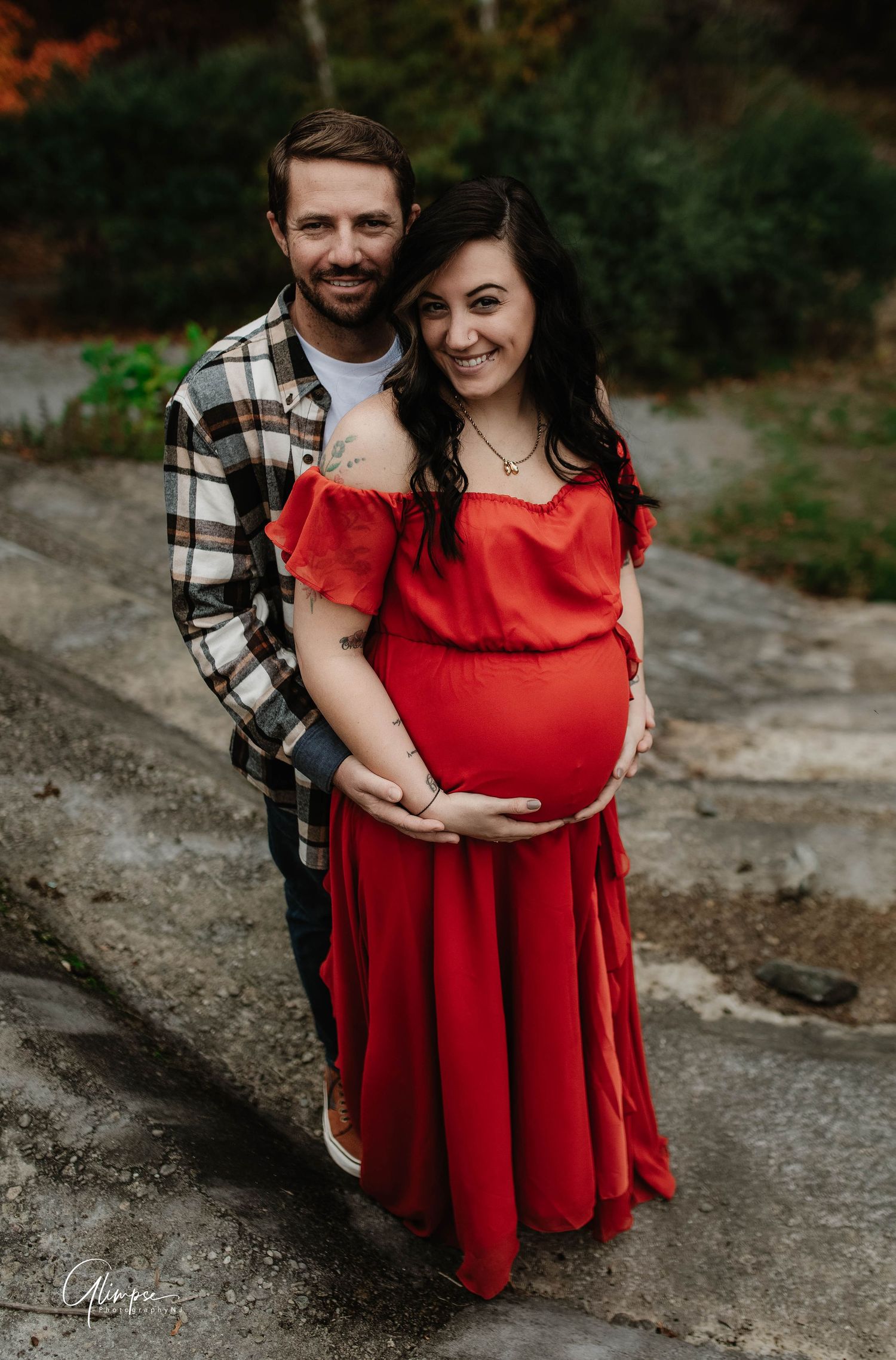 Smiling expectant couple during outdoor maternity shoot, woman in red dress with partner lovingly holding her bump.