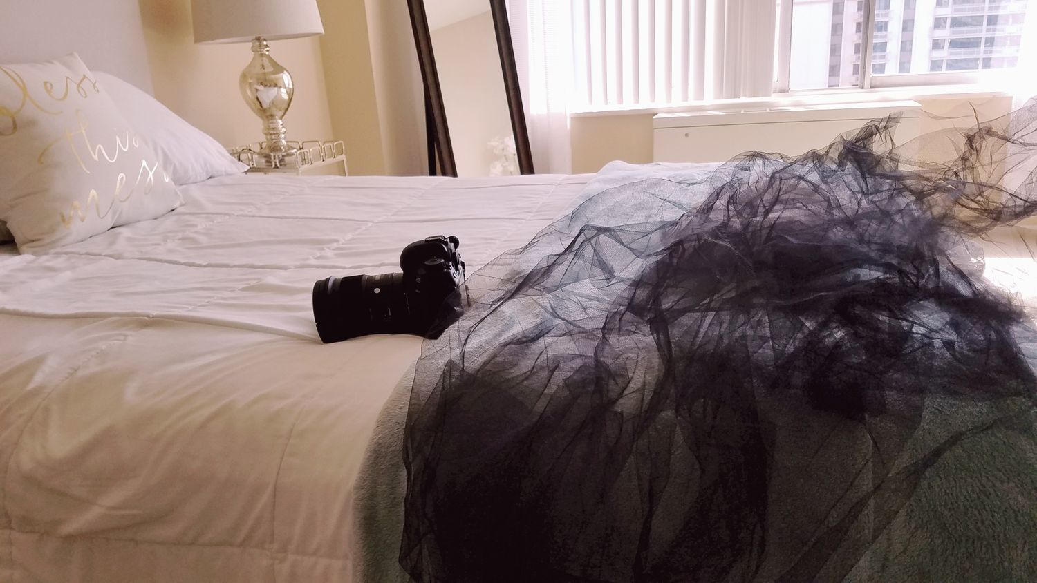 A messy unmade bed with white sheets and dark clothing items scattered on top.