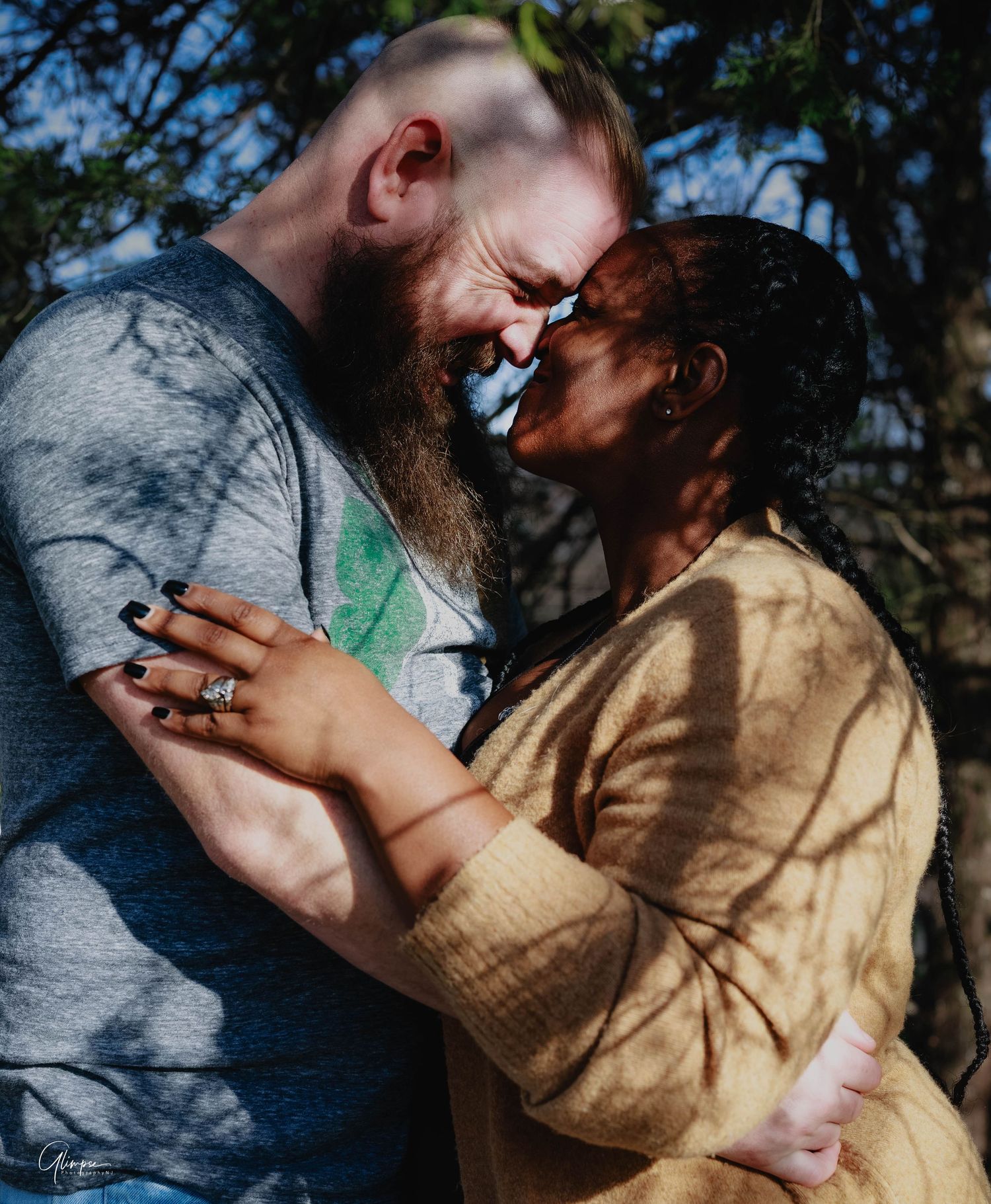 An interracial couple laughs and embraces outdoors in dappled sunlight during an engagement photo session.