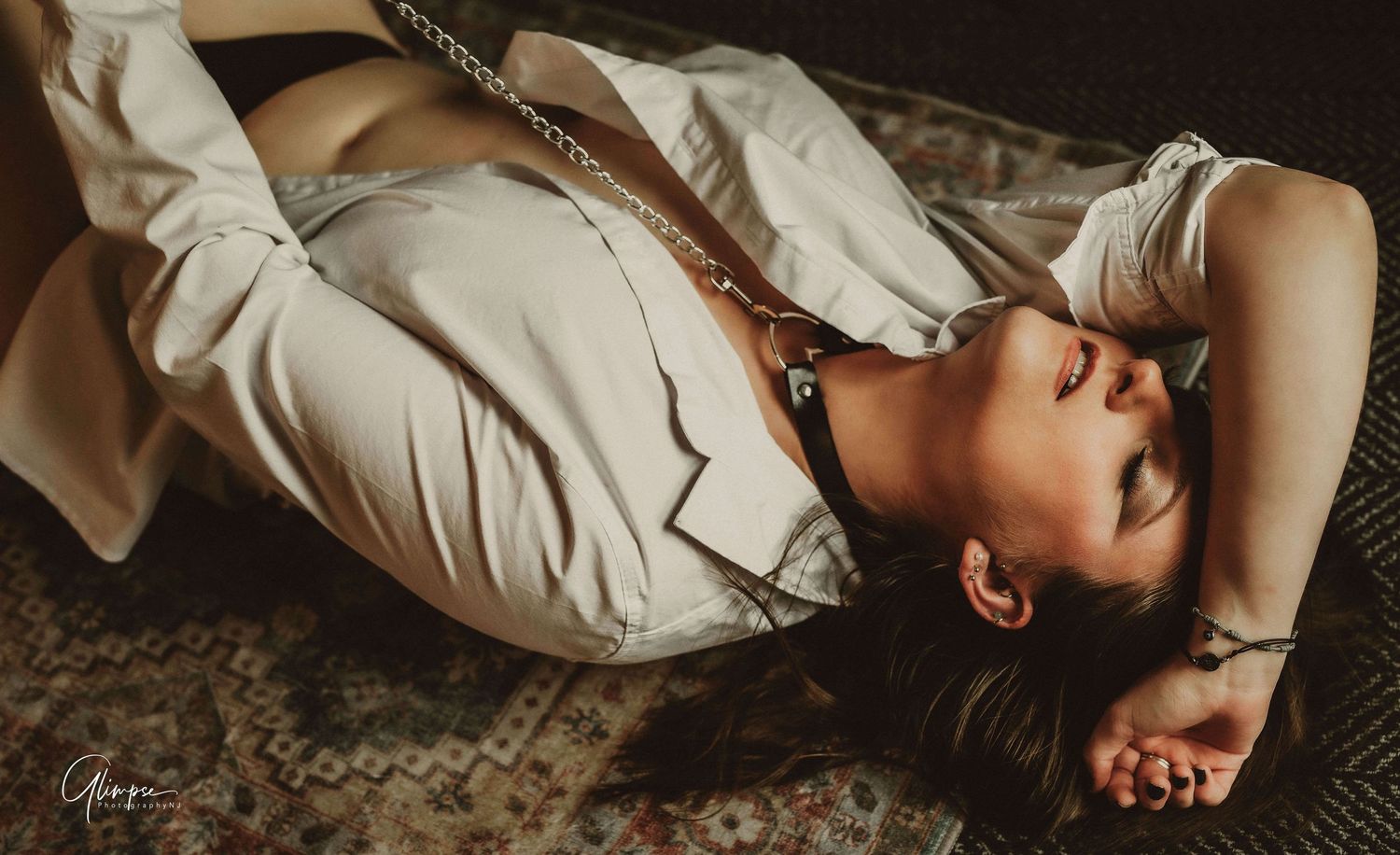Woman in white shirt lying on floor with chain necklace in moody boudoir photography.