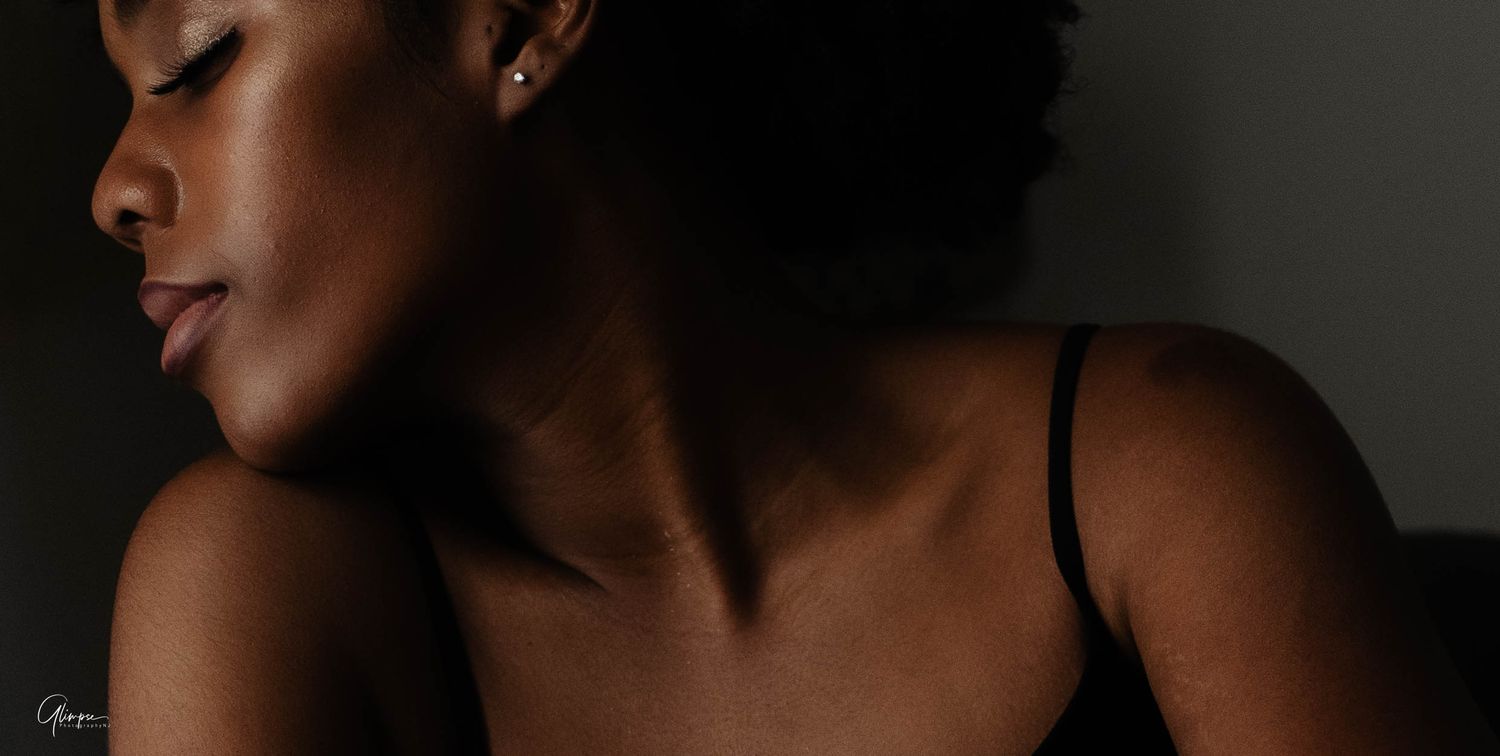 Close-up portrait of a Black woman with natural hair wearing a black bra strap in soft dramatic shadow lighting.