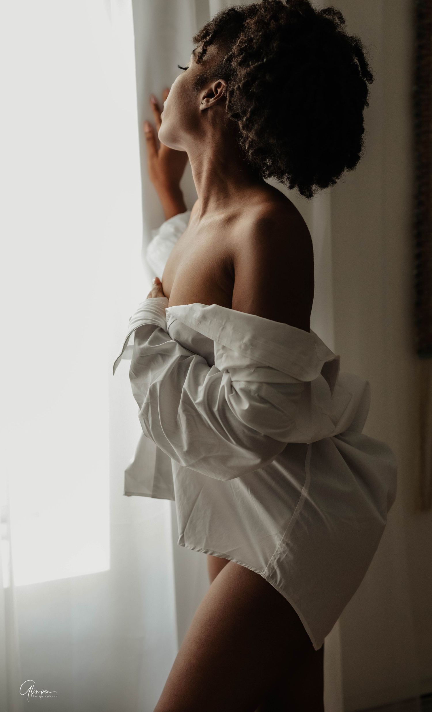 A woman with curly updo poses by a bright window wearing an oversized white button-down shirt off her shoulders.